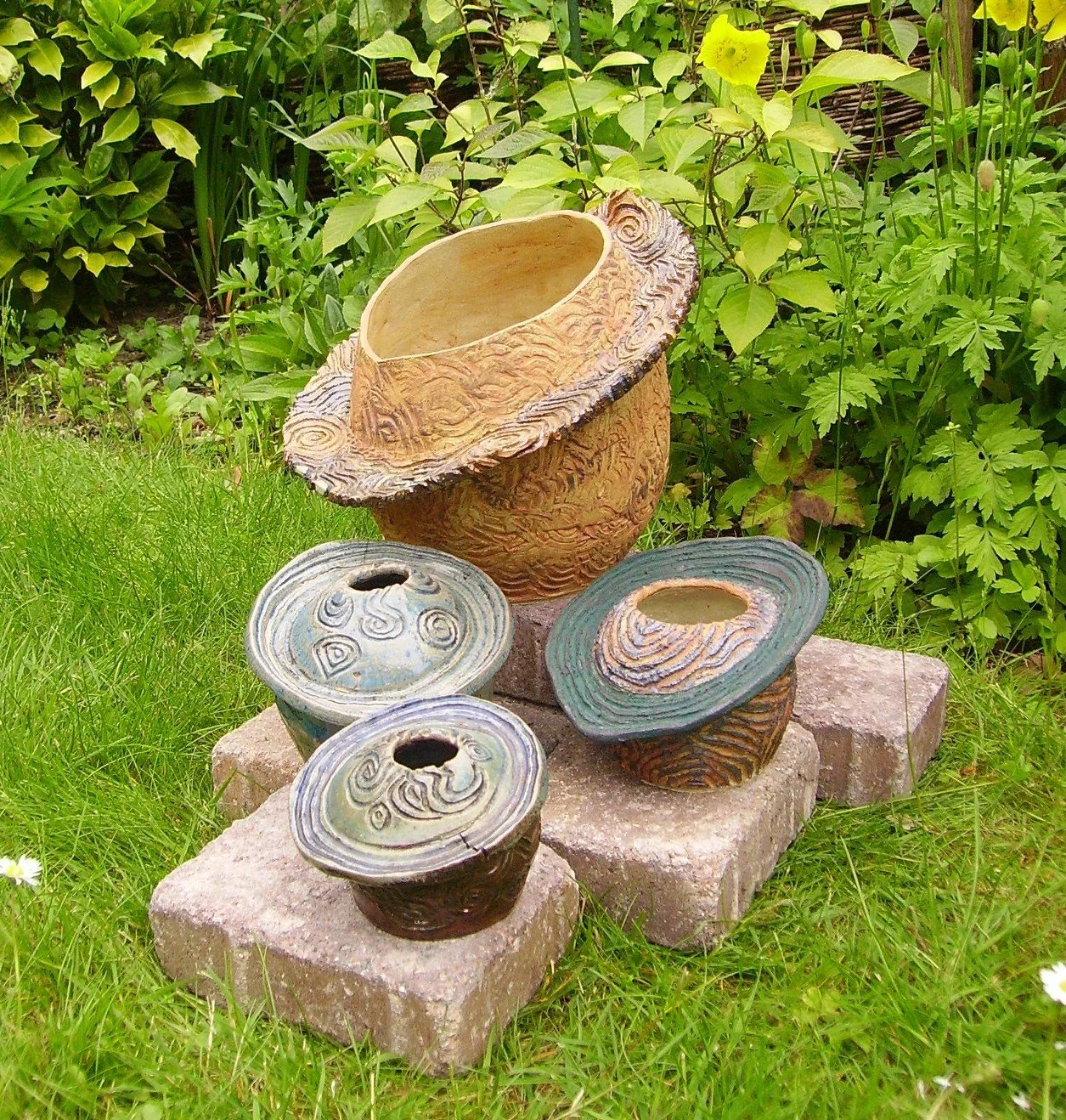 Assorted decorative ceramic pots with textured and colorful designs are arranged on stone bricks in a garden with green plants and grass.