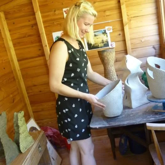 Helen with blonde hair, wearing black is tidying the workshop filled with white pottery inside a wooden-walled room, shed.