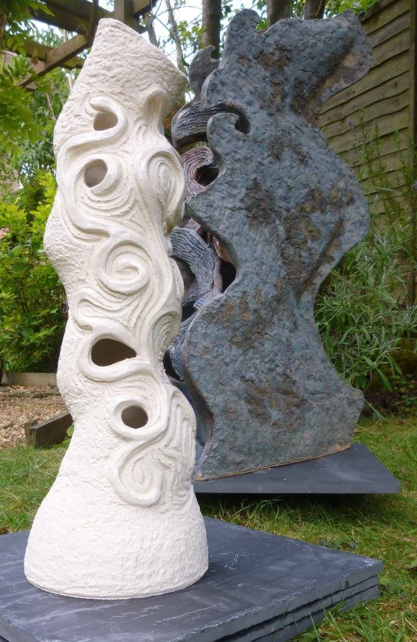 Two abstract stone sculptures with swirling and textured patterns, one in light-coloured clay and the other in dark blue coloured ceramics, displayed outdoors on black bases with greenery and trees in the background.