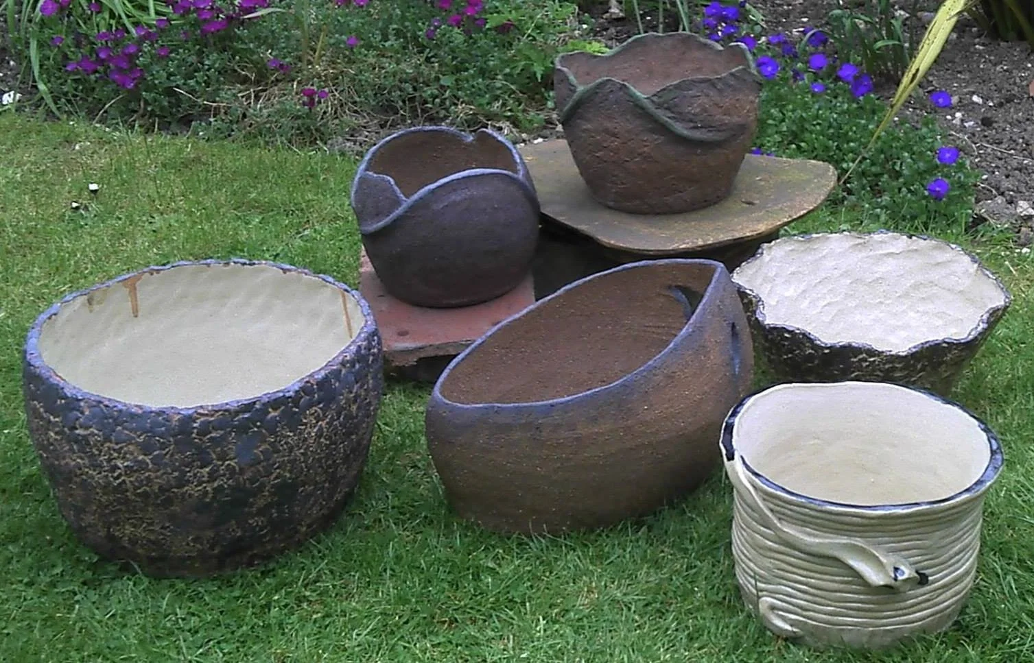 Collection of decorative outdoor ceramic pots with various textures and colors on a grassy lawn, with purple flowers and garden plants in the background.