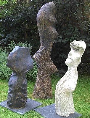 Three abstract stone sculptures outdoors on grass, with bushes in the background.