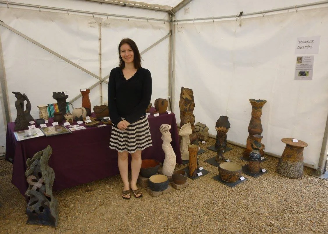 Woman standing inside a tent at an art exhibit, surrounded by various ceramic sculptures and pottery displayed on tables and on the ground.