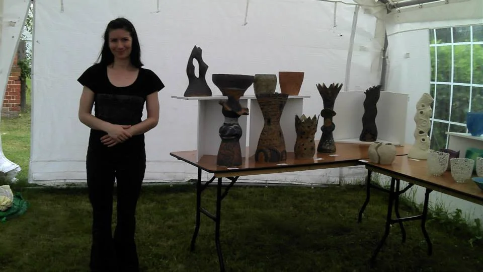 A woman standing inside a white tent next to a display of various ceramic sculptures and pottery on tables.
