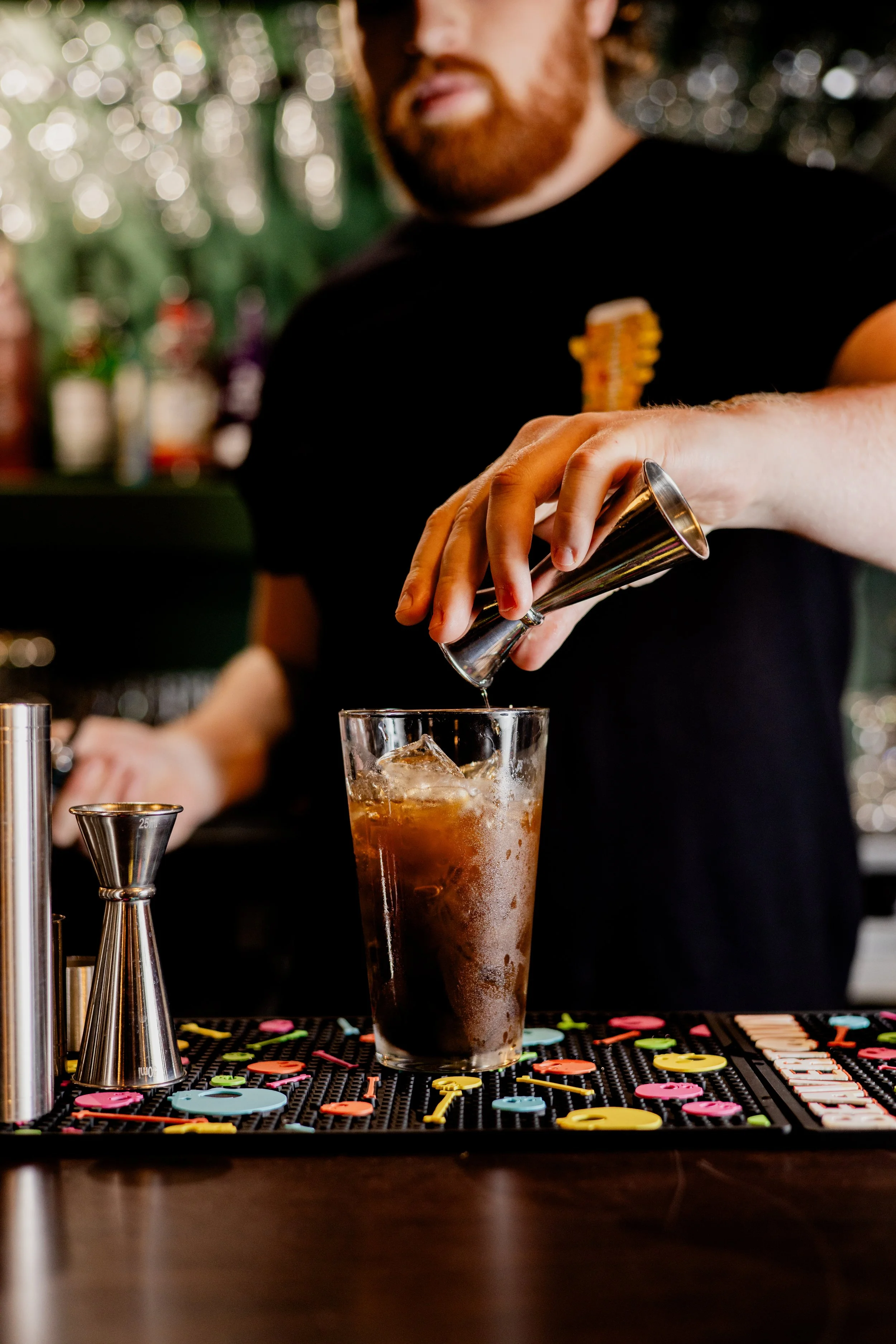 Bartender pouring a drink into a tall glass with ice, on a bar counter decorated with colorful drink stirrers and a jigger nearby.