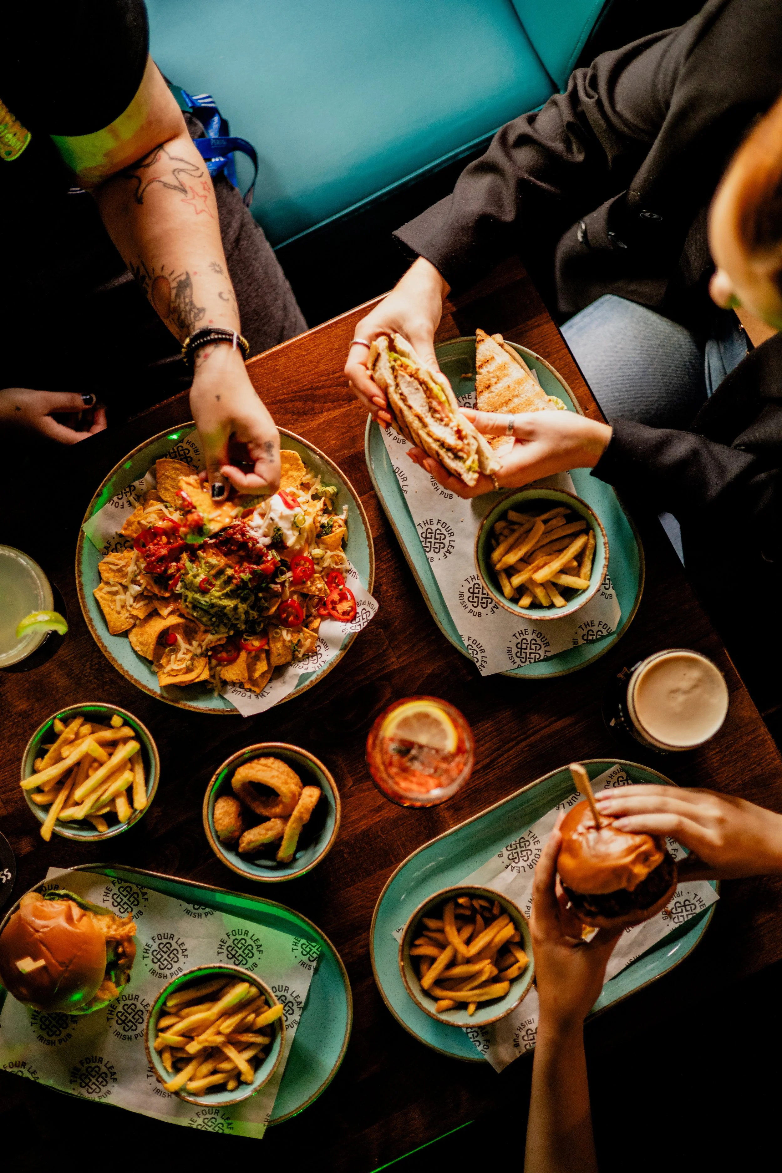 Top-down view of a restaurant table with various dishes including a burger, nachos with toppings, fries, onion rings, and drinks, with people reaching for the food.