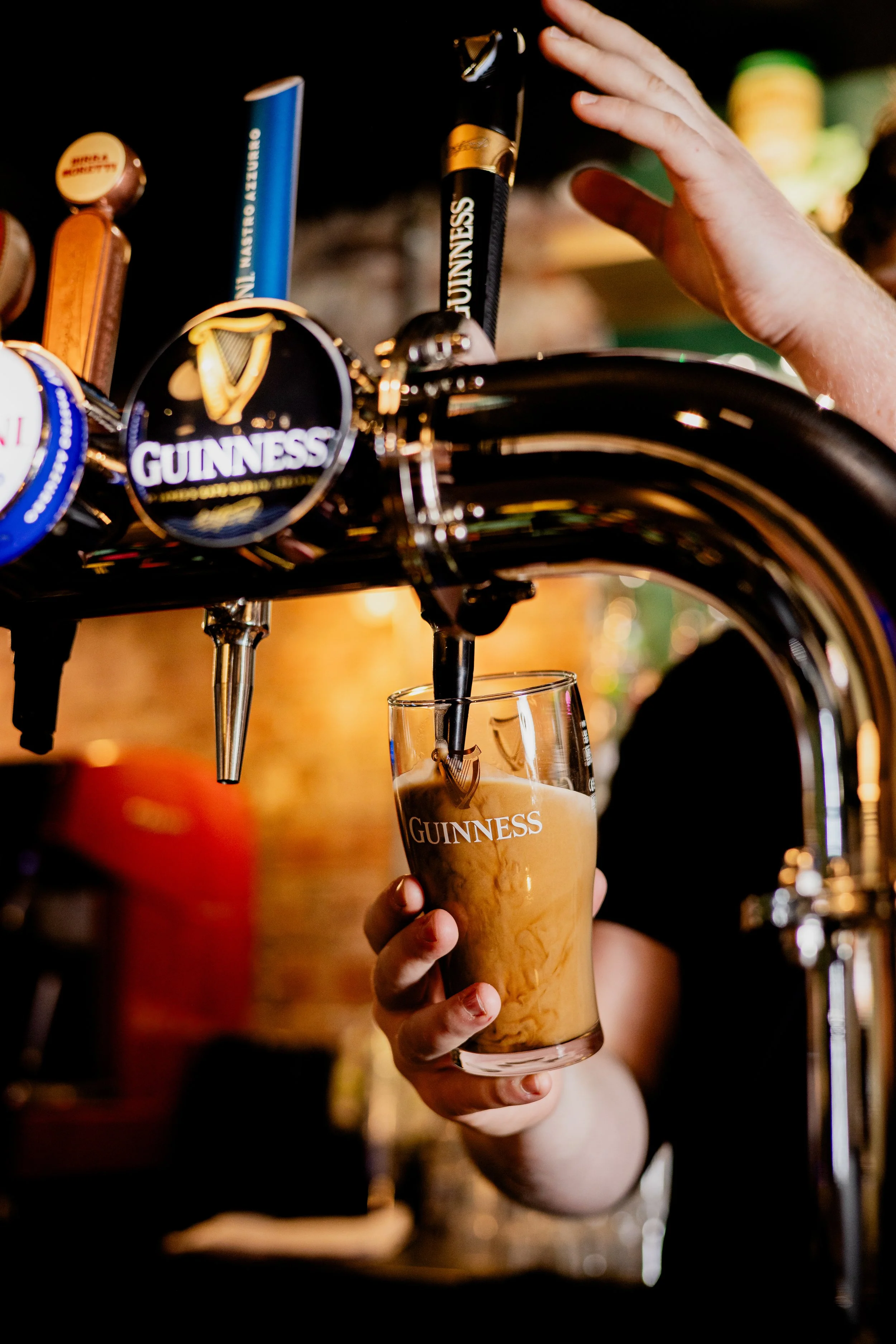 A person holding a glass of Guinness beer underneath a tap, with the beer flowing into the glass. The tap features Guinness branding.