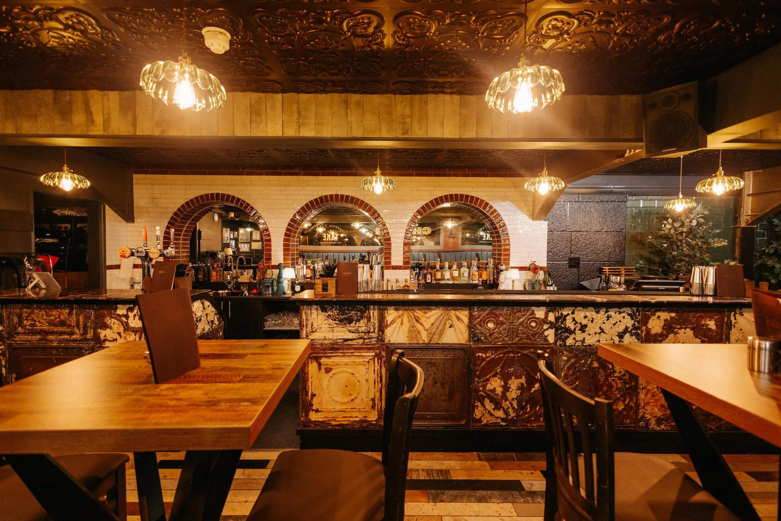 Interior of a cozy restaurant or bar with wooden tables and chairs, ornate pendant lighting, brick arches behind the bar, and warm lighting creating an inviting atmosphere.