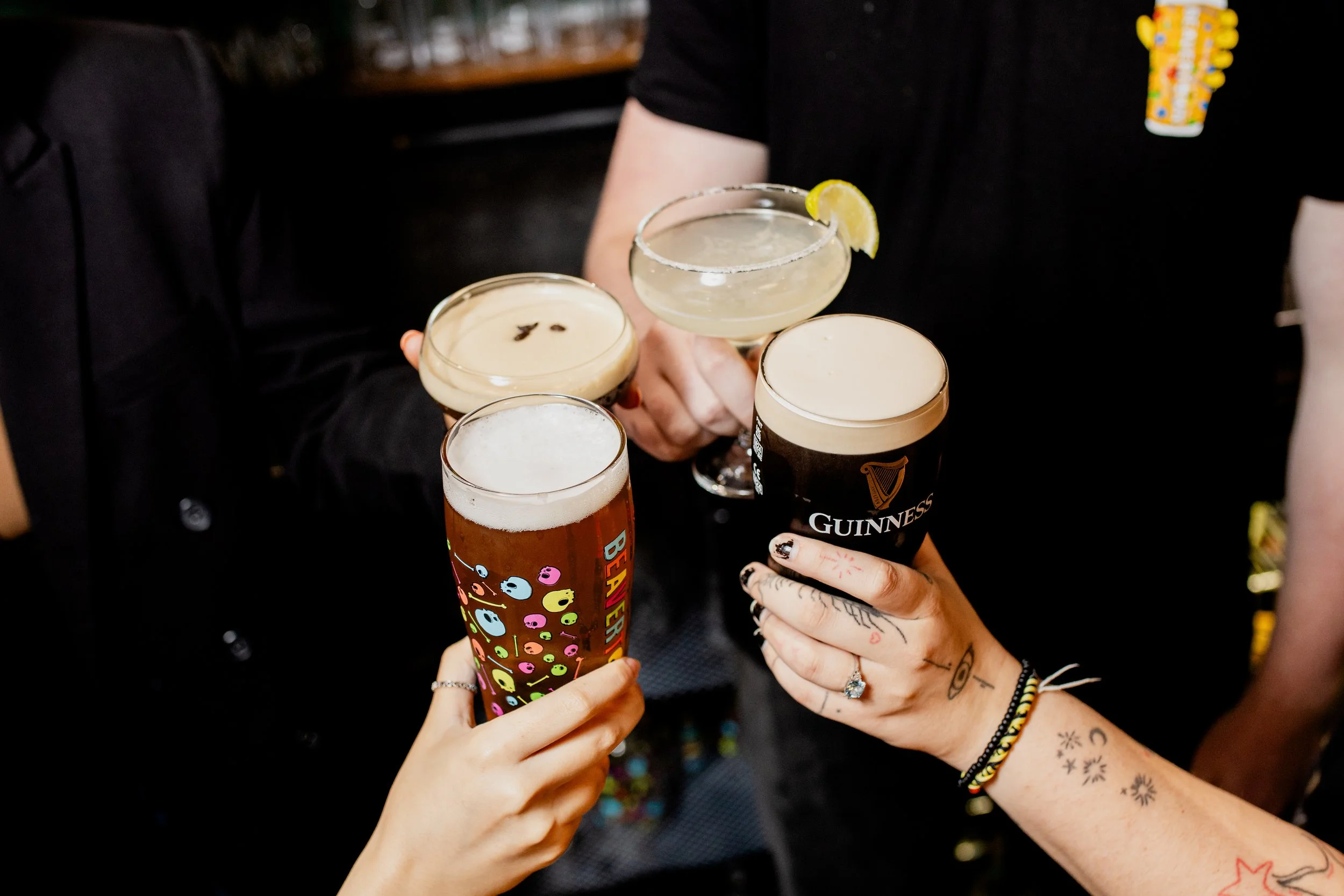 People holding drinks in a bar, including a Guinness beer, a cocktail with a lemon slice and salt rim, and a colorful beverage with writing on the glass.
