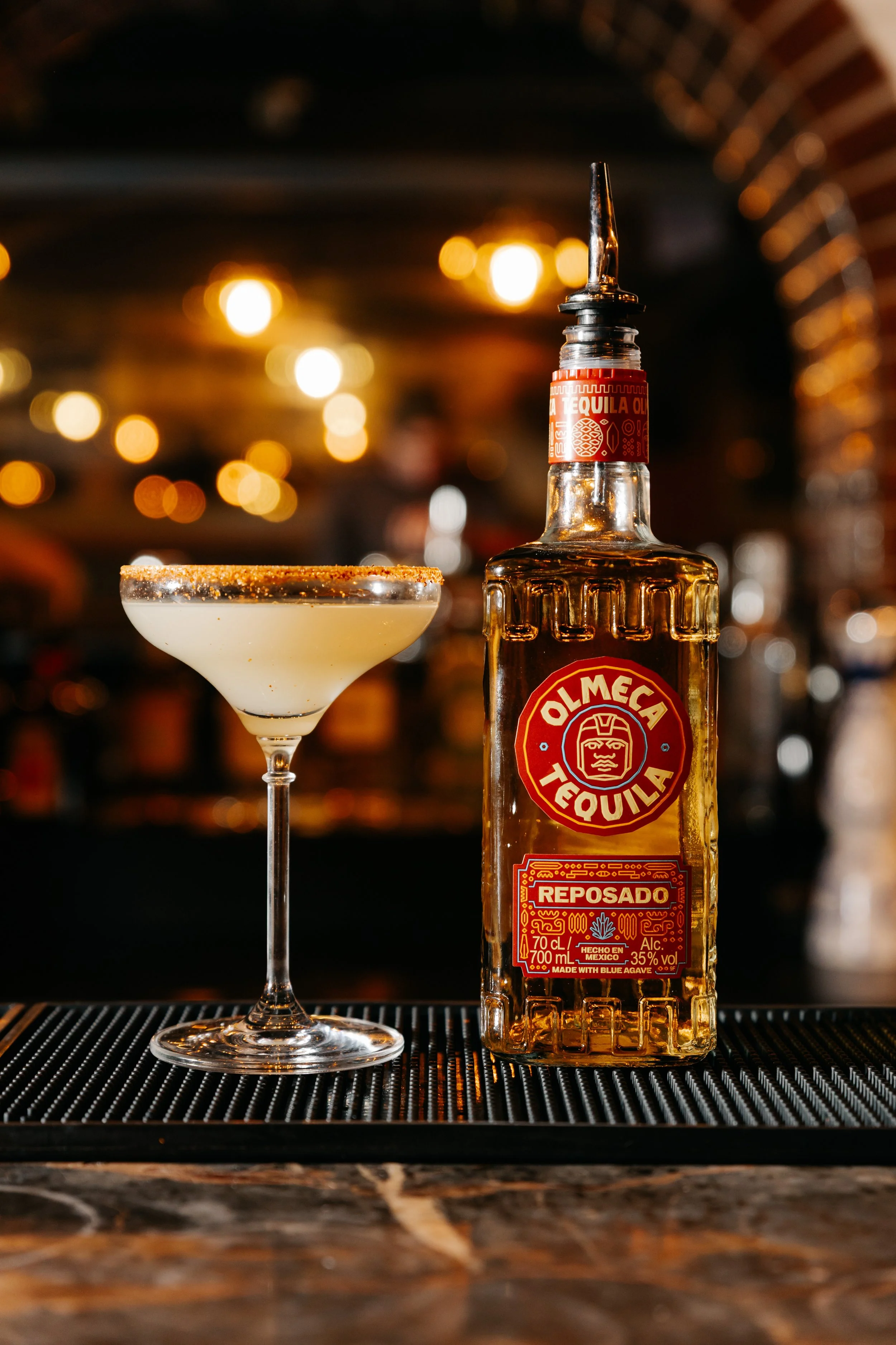 A bottle of Olmeca tequila and a margarita glass with a salted rim, set on a bar counter with warm, blurred bokeh lights in the background.