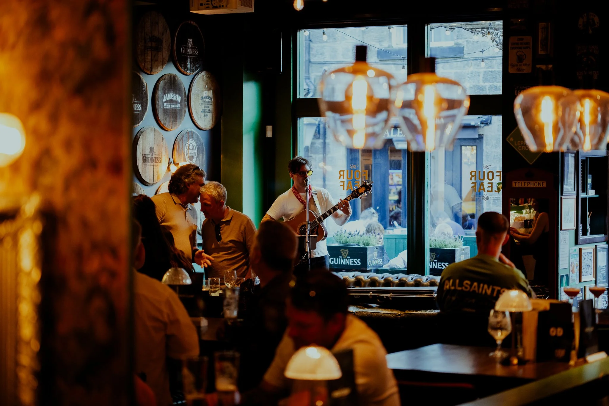 Inside a lively pub with musicians performing on stage and patrons enjoying drinks. The pub has wooden beer barrel decorations on the wall, hanging lights, and a large window showing the street outside.