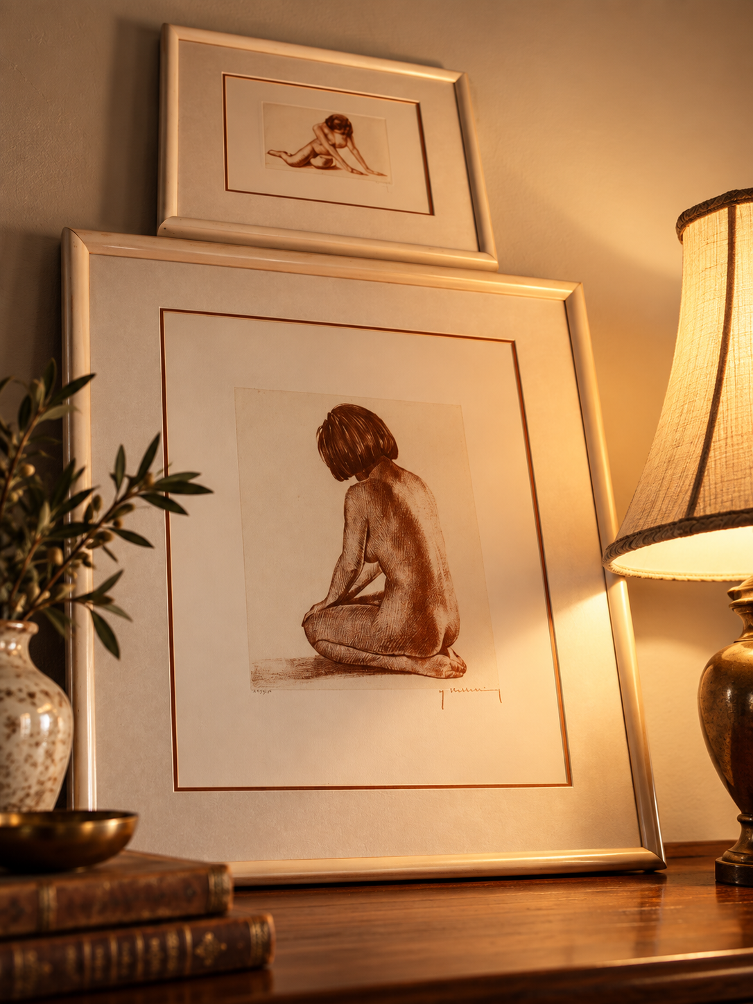 Framed artwork of a nude woman sitting with her back to the viewer, on a wooden table near a lamp and a vase with foliage.