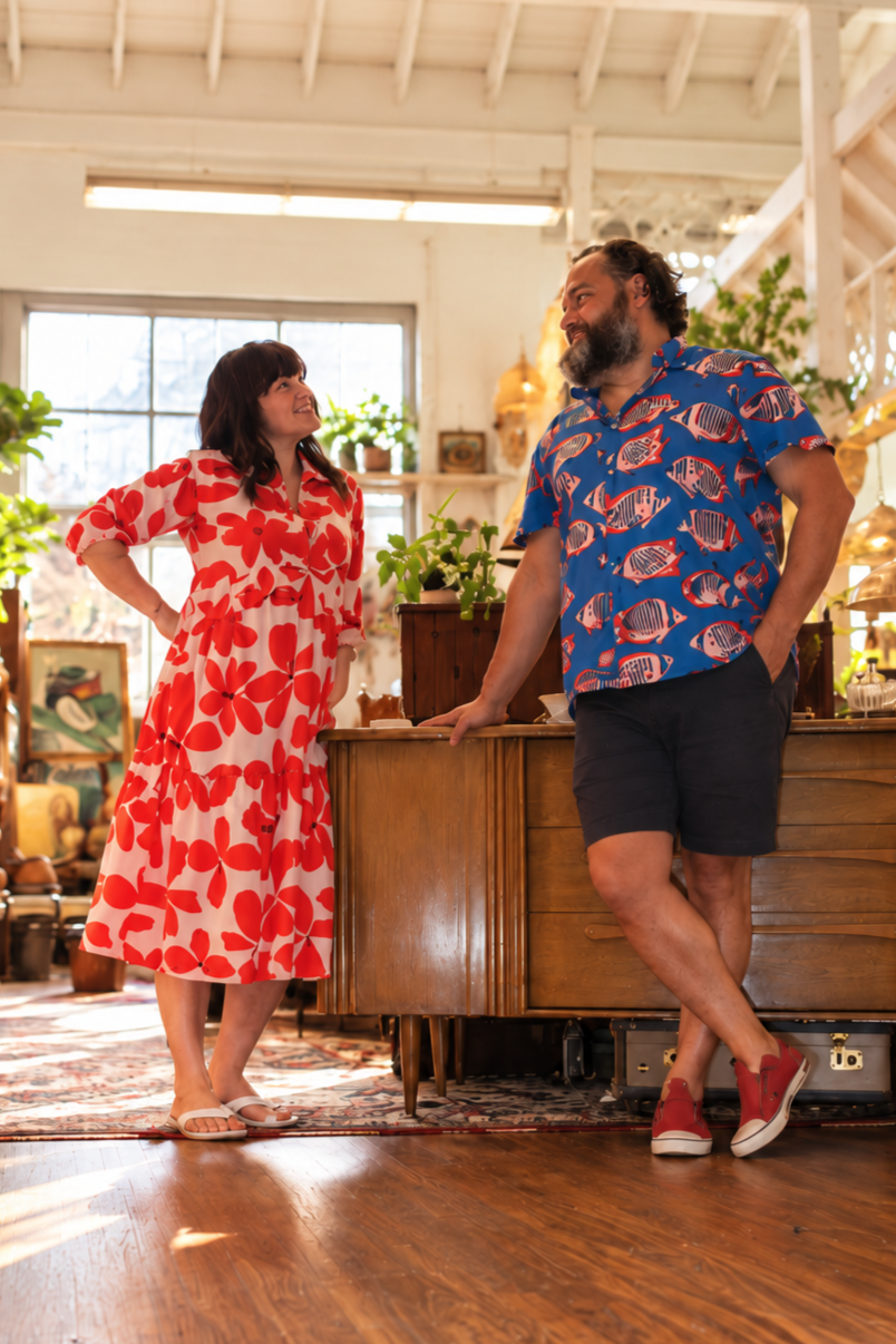 A man and woman are smiling and talking inside an art gallery or shop filled with artworks, plants, and decorative items. The woman wears a red and white floral dress, and the man wears a blue shirt with fish prints and black shorts. They are leaning casually against a wooden counter.