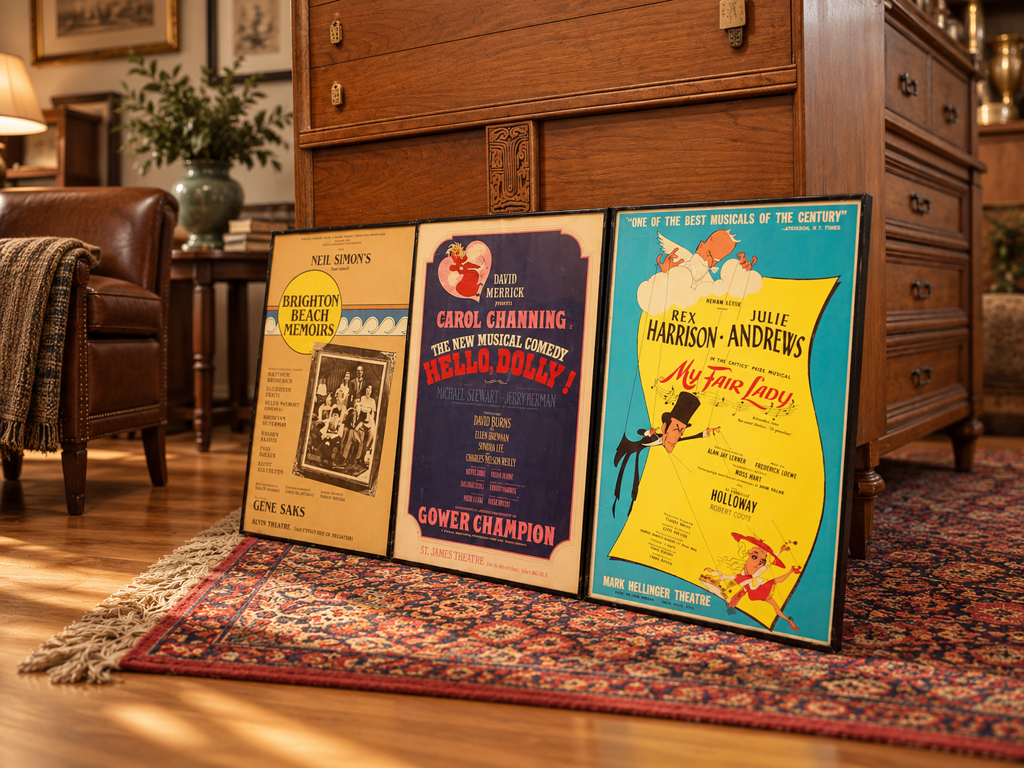 Three vintage musical theater posters resting against a wood cabinet in a cozy, well-lit living room, with a leather armchair, a colorful area rug, and a potted plant in the background.