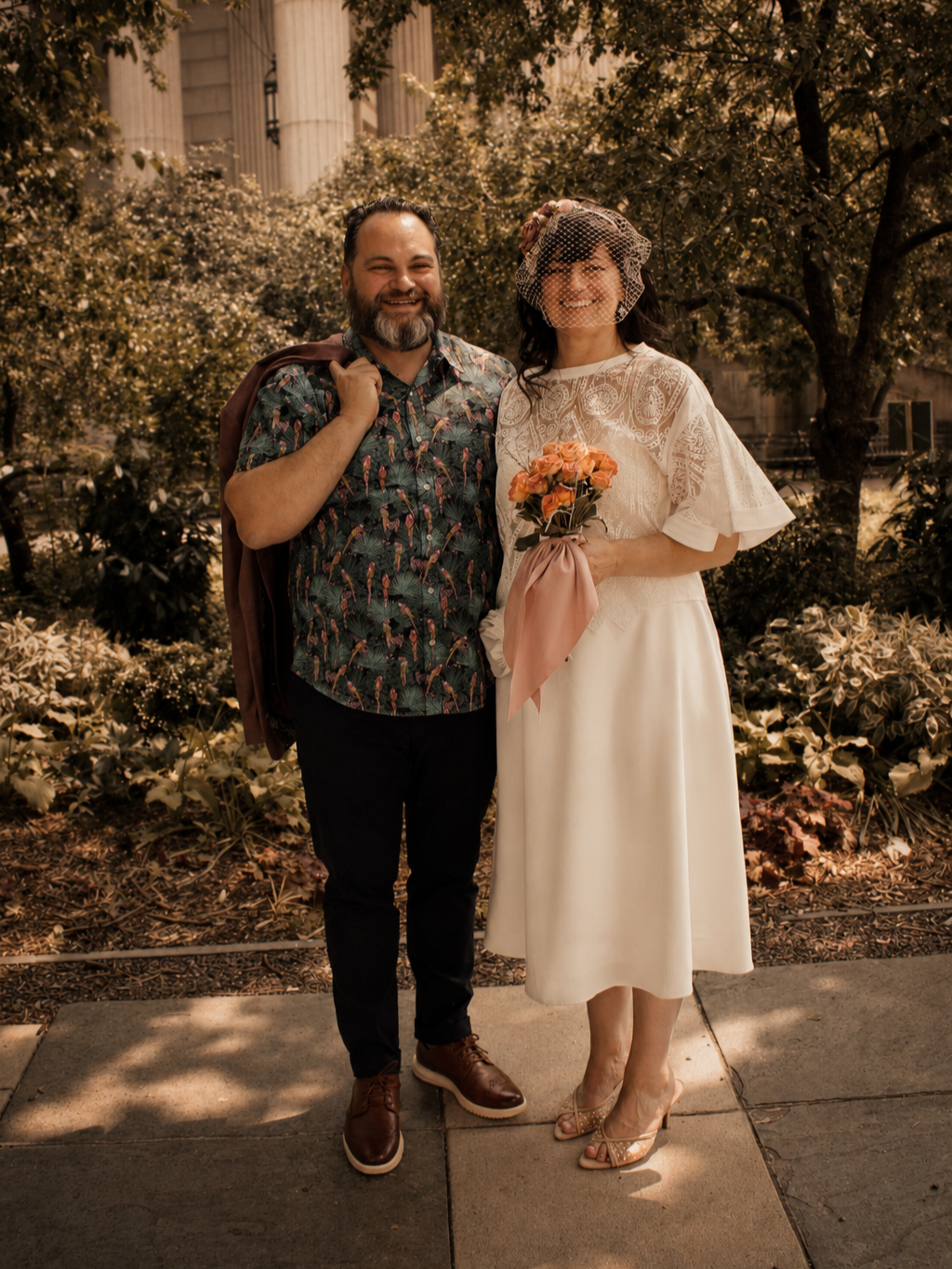 A smiling couple stands outdoors surrounded by trees and plants. The woman is dressed in vintage wedding attire with a lace dress and a birdcage veil, holding a bouquet of peach-colored roses. The man is wearing a patterned shirt and dark pants, with a jacket draped over his shoulder.