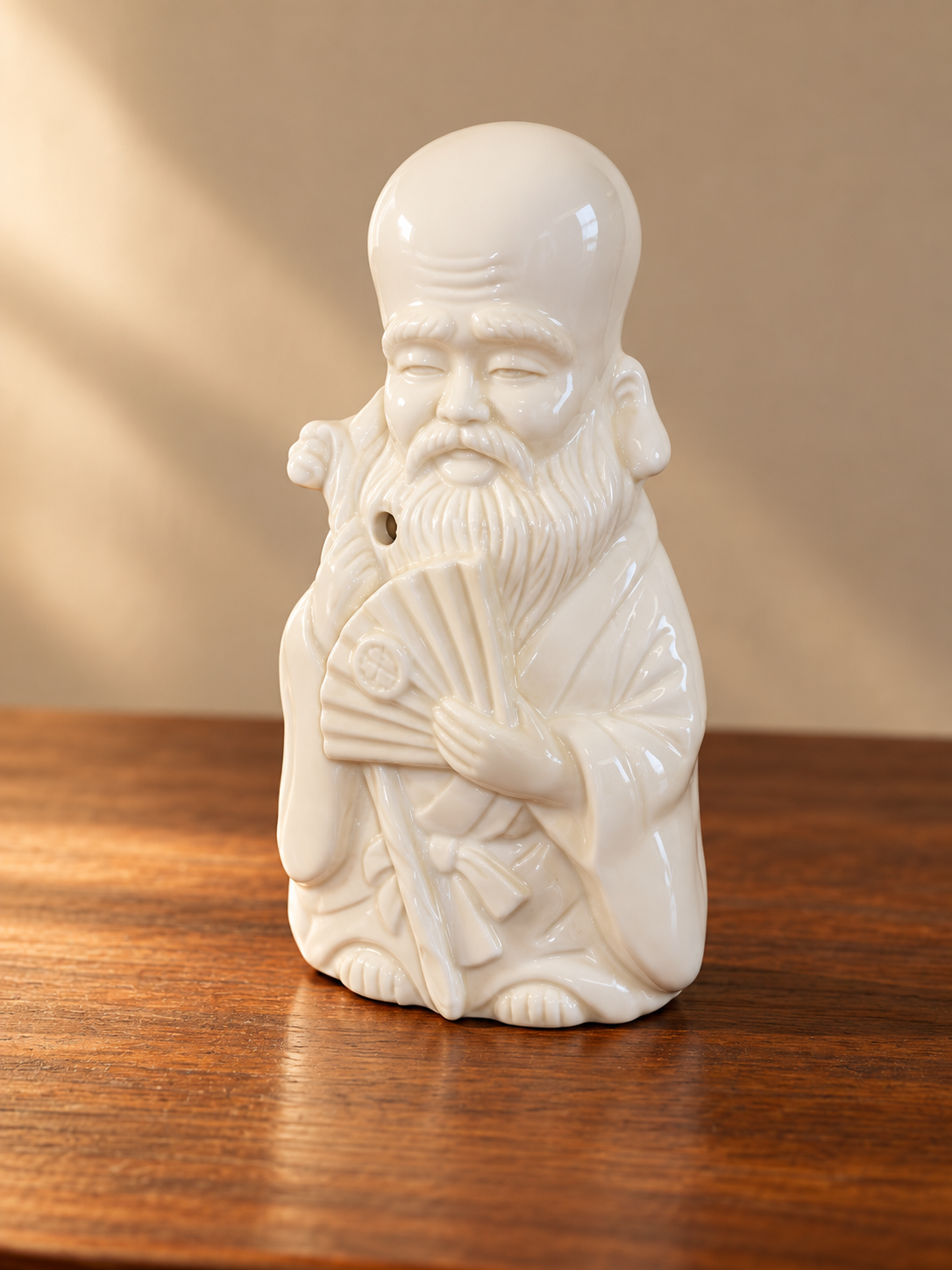 White ceramic figurine of a wise man with a long beard, bald head, holding a fan, standing on a wooden surface.