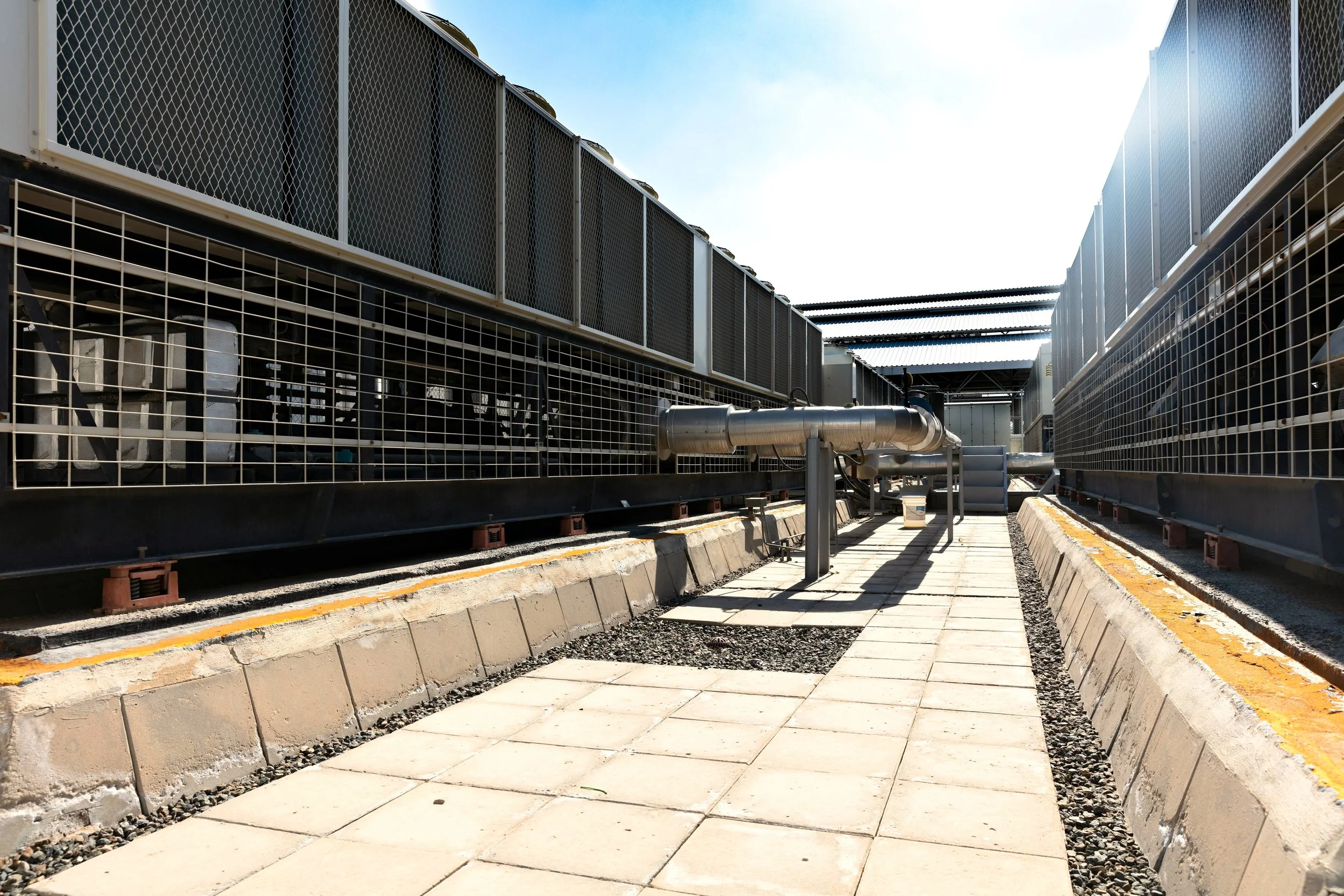 View of industrial cooling or HVAC units enclosed by metal fences, with pipes and ventilation systems, under a clear blue sky.