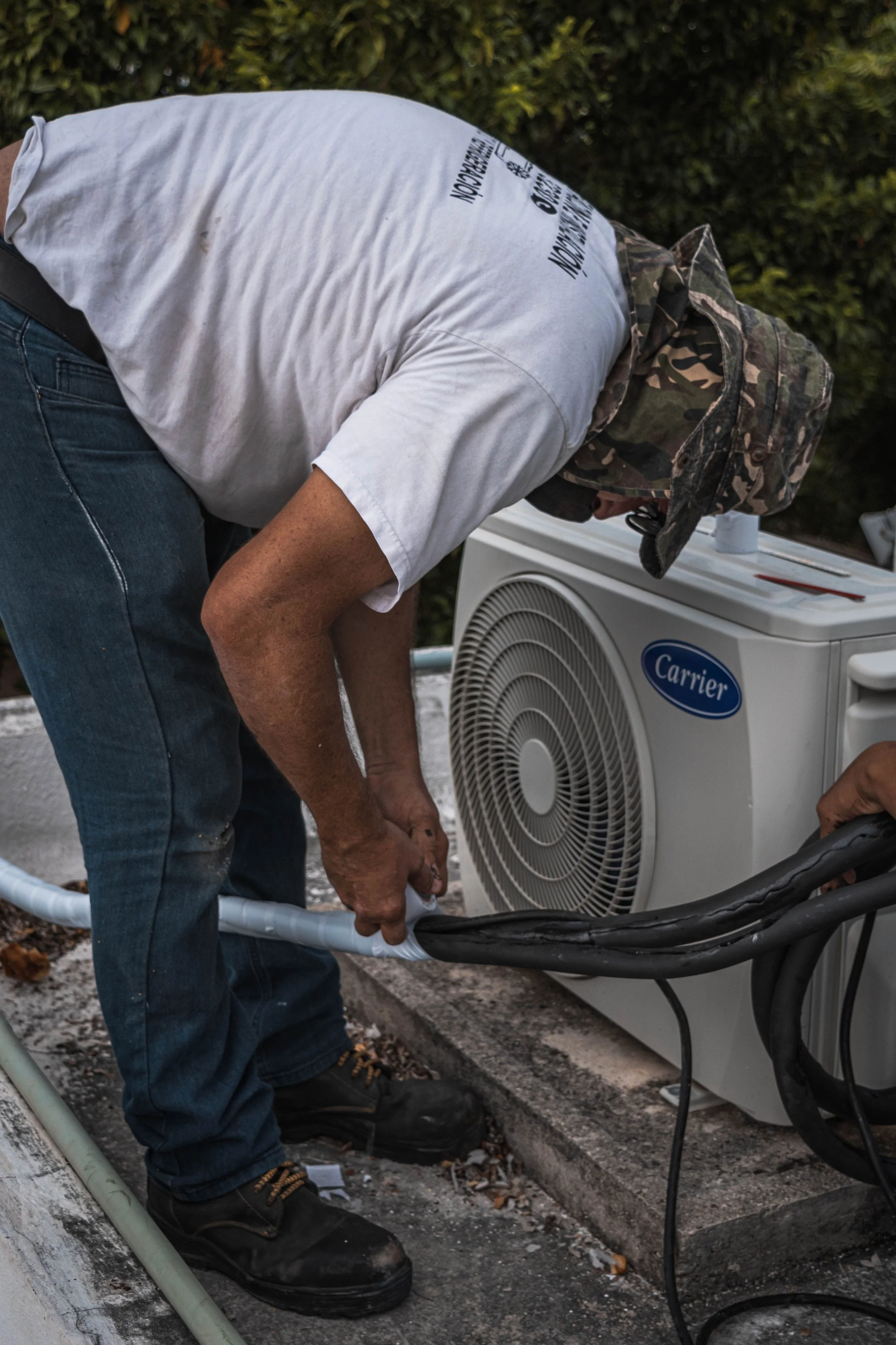 A person wearing a camouflage hat and a white shirt is working outside next to an air conditioning unit on a concrete surface, with a hedge in the background.