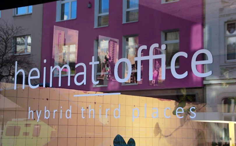 Fensteraussicht auf ein Büro mit Pflanzen und Computern, das den Text 'heimat office' und 'hybrid third places' auf dem Glas zeigt.