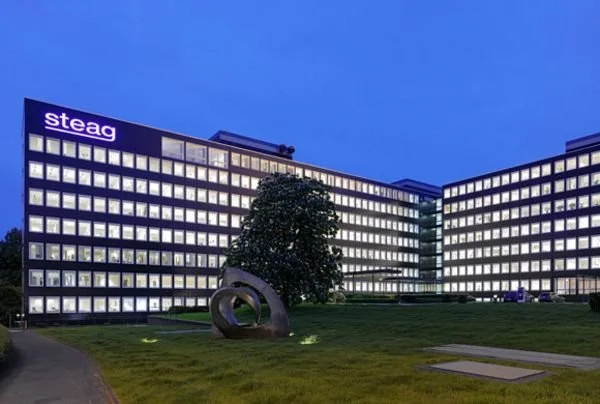 Bild eines großen, mehrstöckigen Bürogebäudes mit dem Logo 'steag' an der Spitze, beleuchtete Fenster und eine Skulptur im Gras vor dem Gebäude