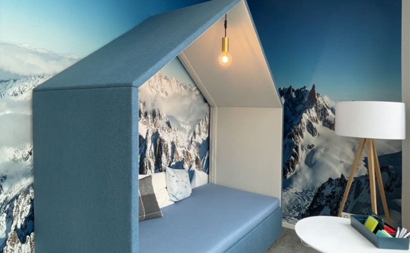 Ein modernes Zimmer mit einer blauen Sitznische, die mit Bergfotografien dekoriert ist. Es gibt eine Stehlampe, einen kleinen Tisch mit Stiften und einen schwarzen Hocker auf einem braunen Fellteppich.