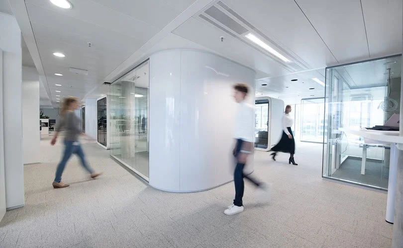 Moderne Büroetage mit weißen Wänden, gläsernen Abtrennungen und mehreren Personen, die durch den Raum gehen.