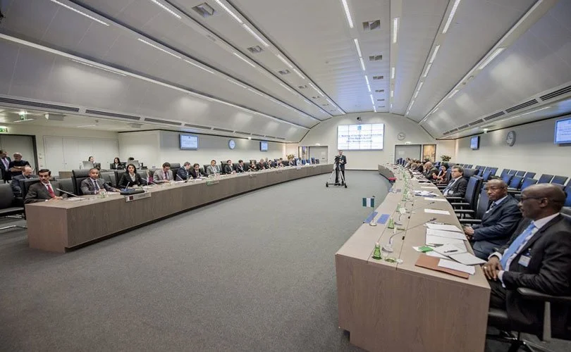 Ein großes, modernes Konferenzzimmer mit Teilnehmenden, die um einen langen runden Tisch sitzen, viele Monitore an den Wänden, ein Mann auf einem Roller in der Mitte, die meisten Personen tragen Anzüge.
