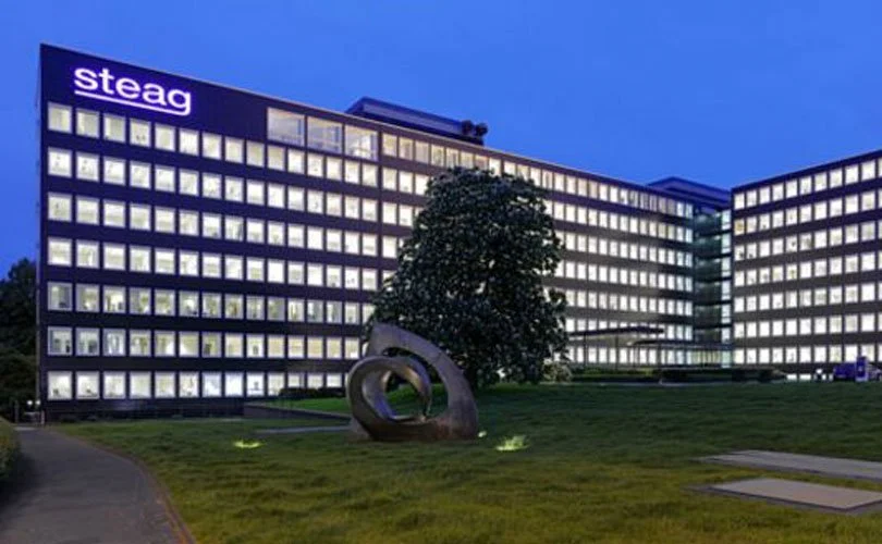 Bild eines großen, mehrstöckigen Bürogebäudes mit dem Logo 'steag' an der Spitze, beleuchtete Fenster und eine Skulptur im Gras vor dem Gebäude