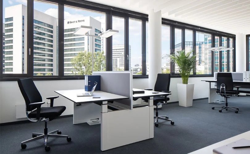 Modernes Büro mit großen Fenstern, Schreibtischen, schwarzen Drehstühlen, einem Laptop, Aktenordnern, einer Pflanze und Blick auf Hochhäuser im Stadtzentrum.