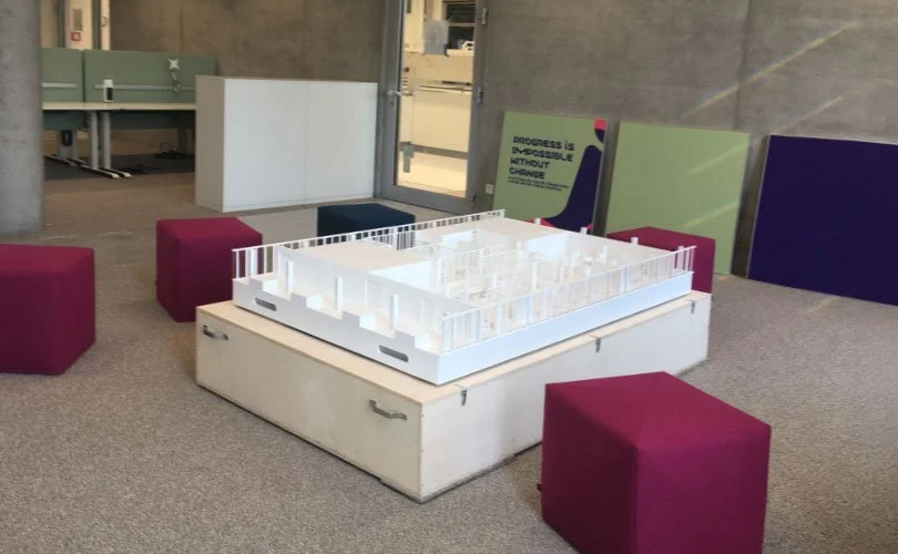 Innenraum eines modernen Gebäudes mit Sitzgelegenheiten, einem Tisch mit einem Modell, sowie Schildern und einem Fenster. Es sind Sitzwürfel in Magenta, ein Modell eines Gebäudes und Poster an der Wand zu sehen.
