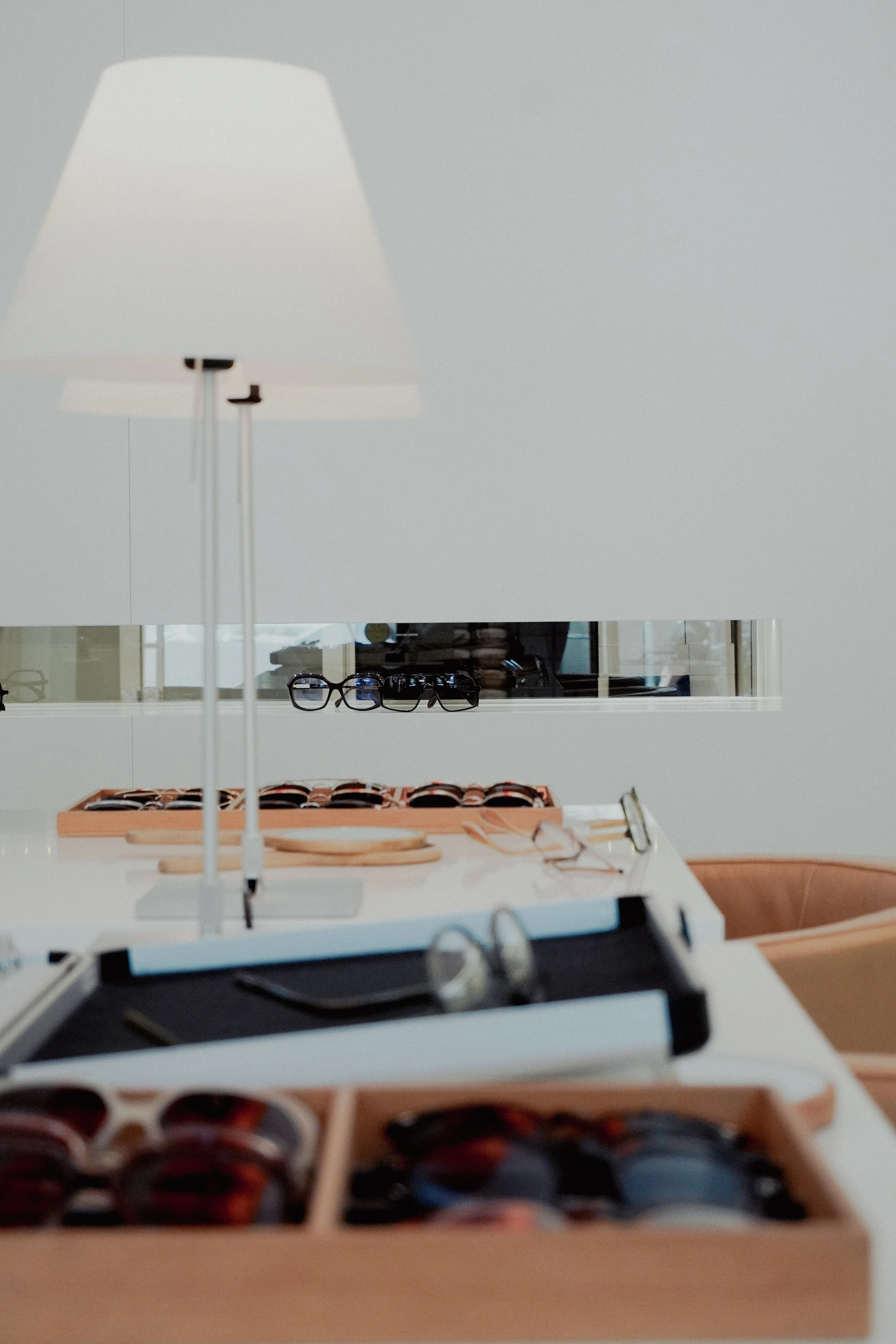 Eine Brille, Sonnenbrillen, auf einem weißen Tisch in einem Optikergeschäft, im Hintergrund ein Regal mit Sonnenbrillen und optischen Brillen. Ein Lampenschirm ist im Vordergrund sichtbar.