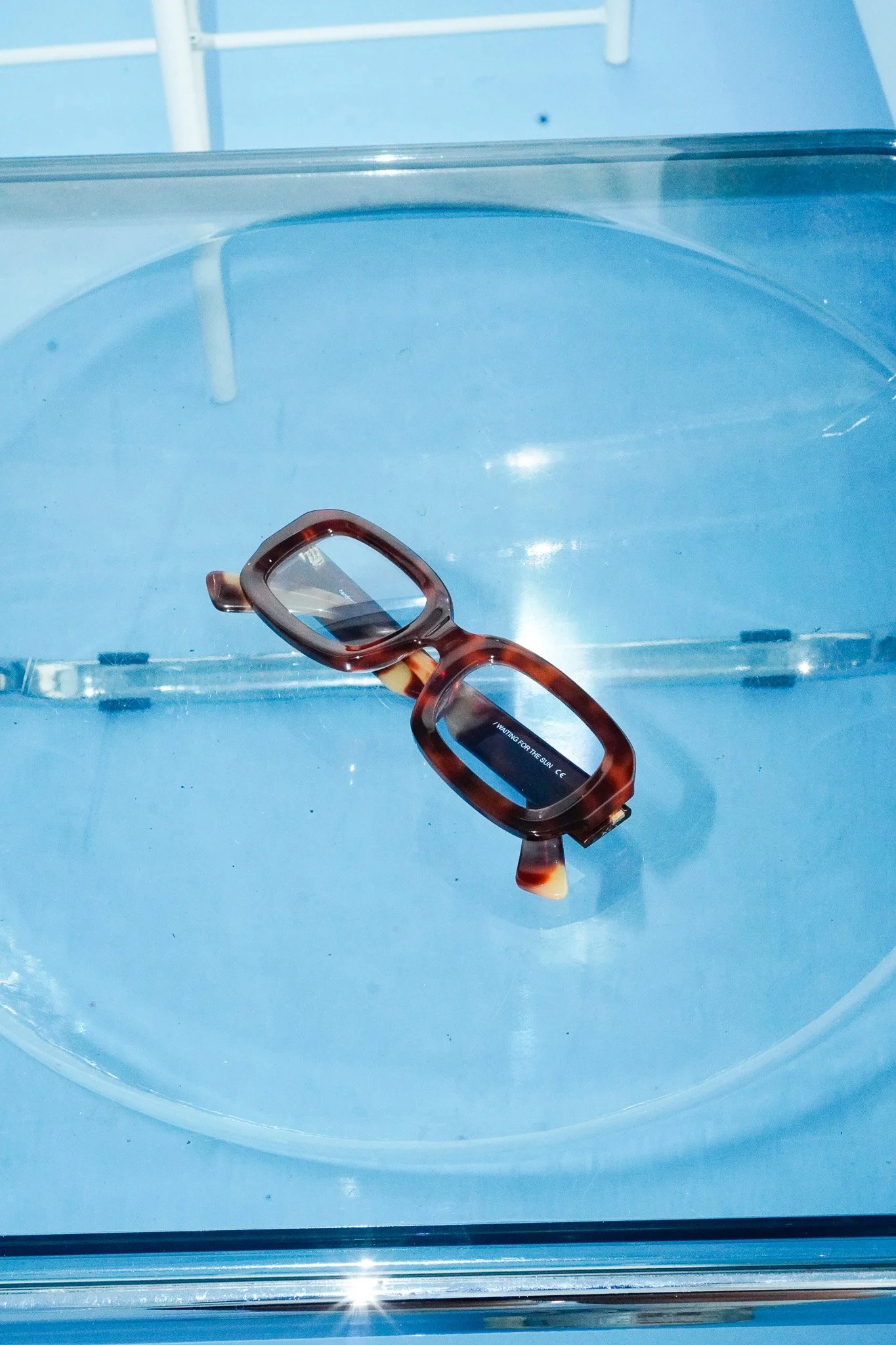 Brille liegt auf einer transparenten Oberfläche, im Hintergrund blaue Wand mit weißen Elementen.