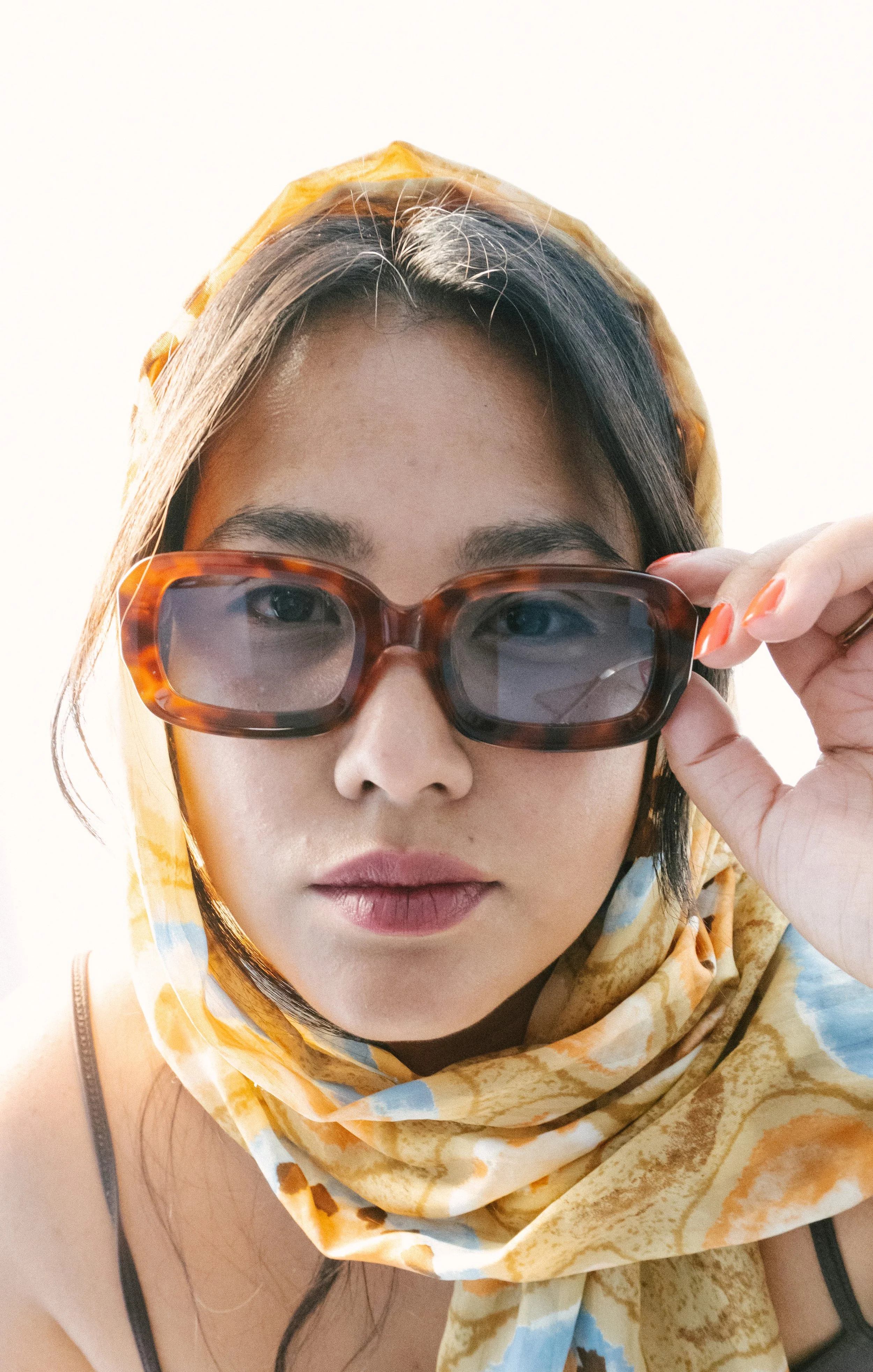Nahaufnahme eines jungen Frauen, die große, braune Sonnenbrille trägt, eine gelbe Latzhose und einen gelben Kopftuch mit blauen Mustern trägt, während sie die Brille anhebt.