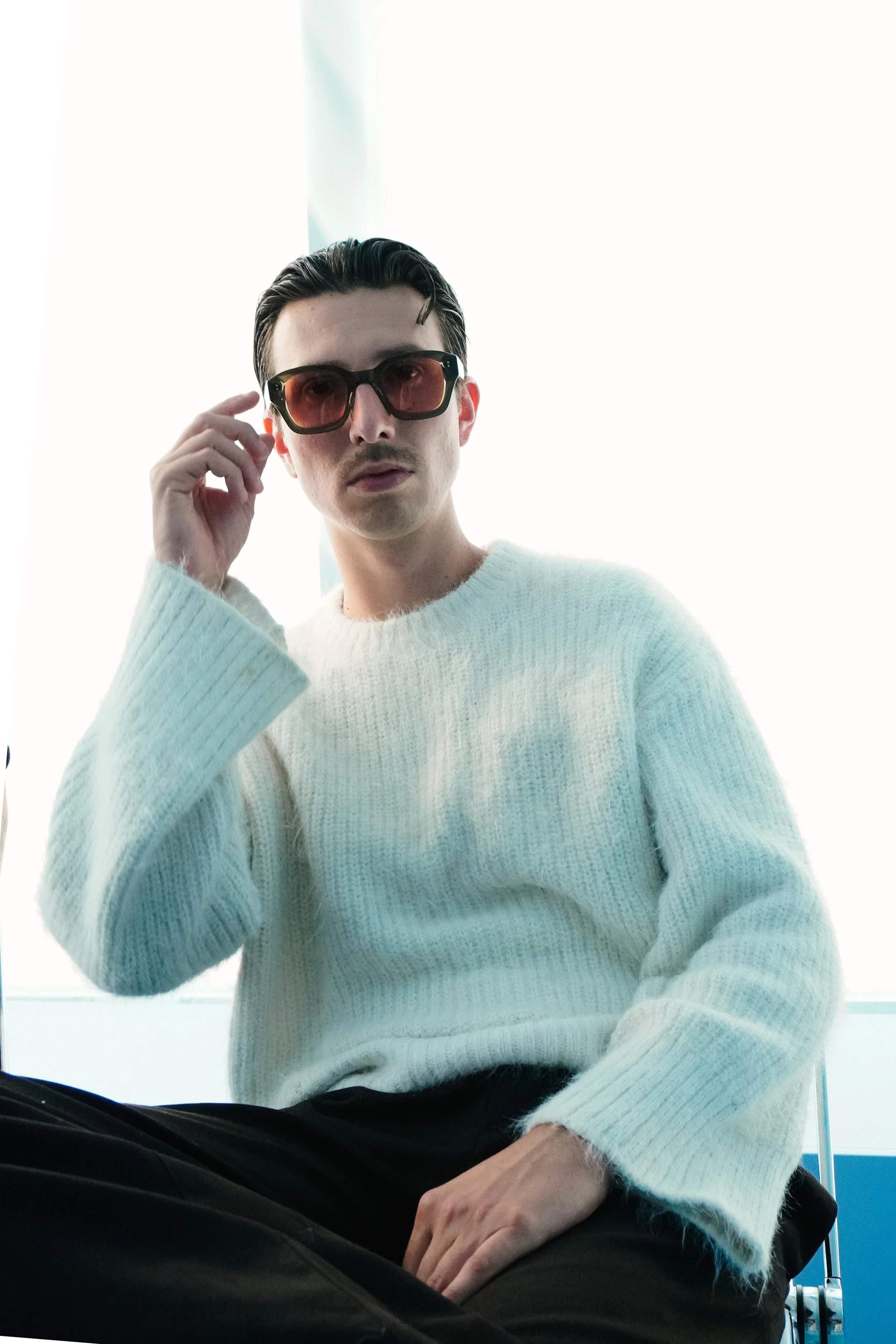 Junge Person mit dunklen Haaren, trägt runde Sonnenbrille mit rotem Tint auf einem weißen Pullover, sitzt in einem Flugzeug oder Zug, hoher Fenster im Hintergrund.
