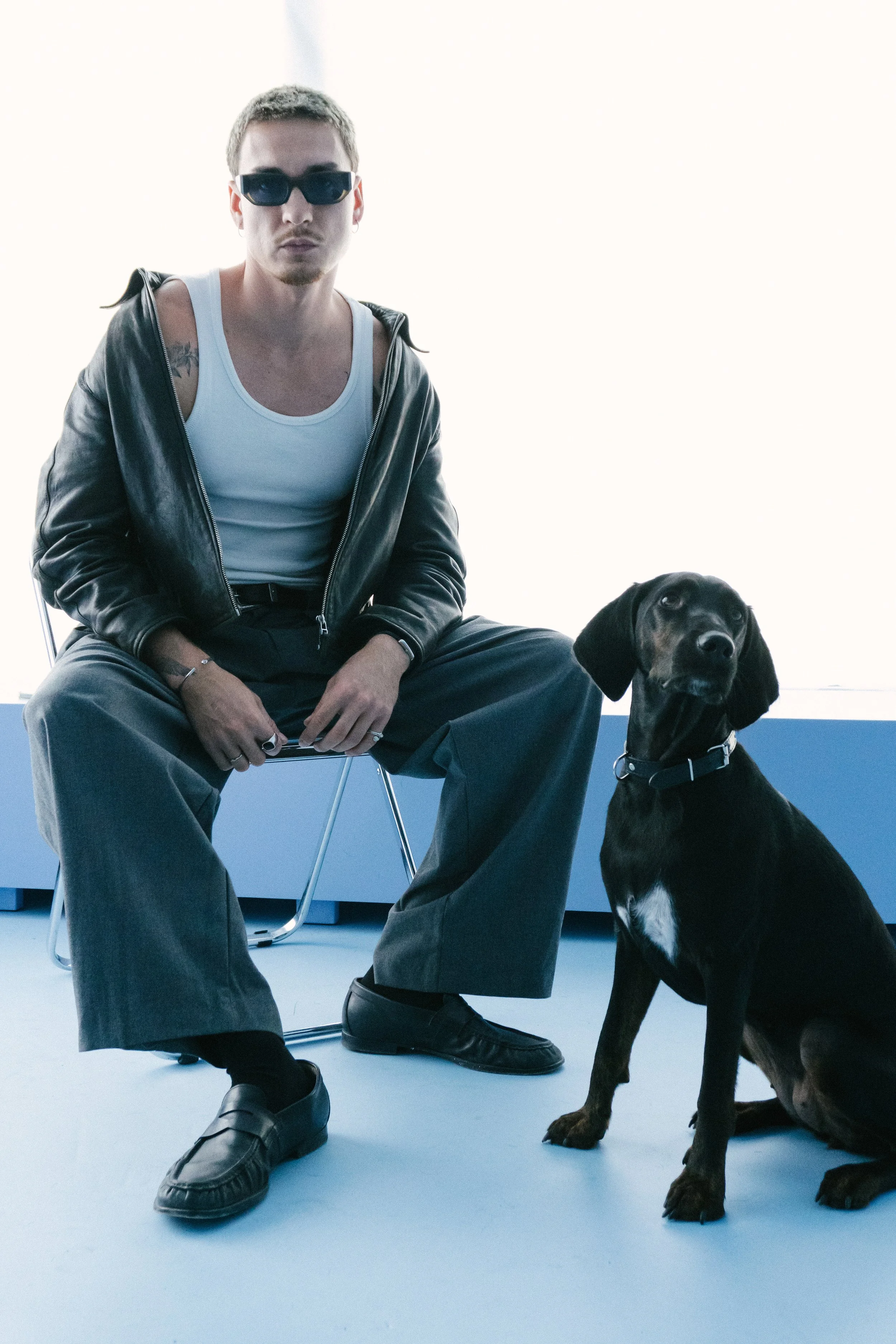 Ein junger Mann trägt dunkle Sonnenbrille, weiße Tanktop, schwarze Lederjacke und Jeans, sitzt auf einem Stuhl, neben ihm sitzt ein schwarzer Hund mit braunen Pfoten, vor einem hellen Hintergrund.