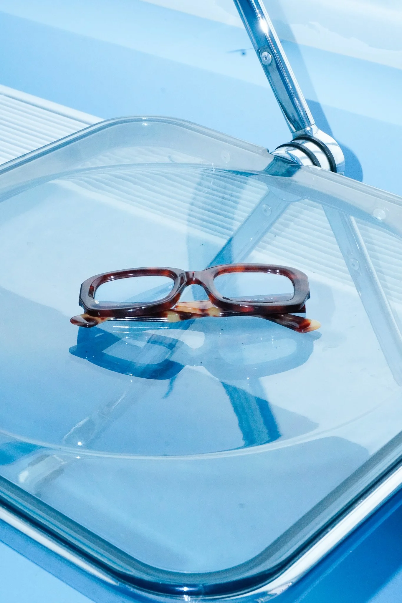 Brille liegt auf einer runden, transparenten Glasplatte, im Hintergrund eine moderne Leuchte und blauer Himmel.