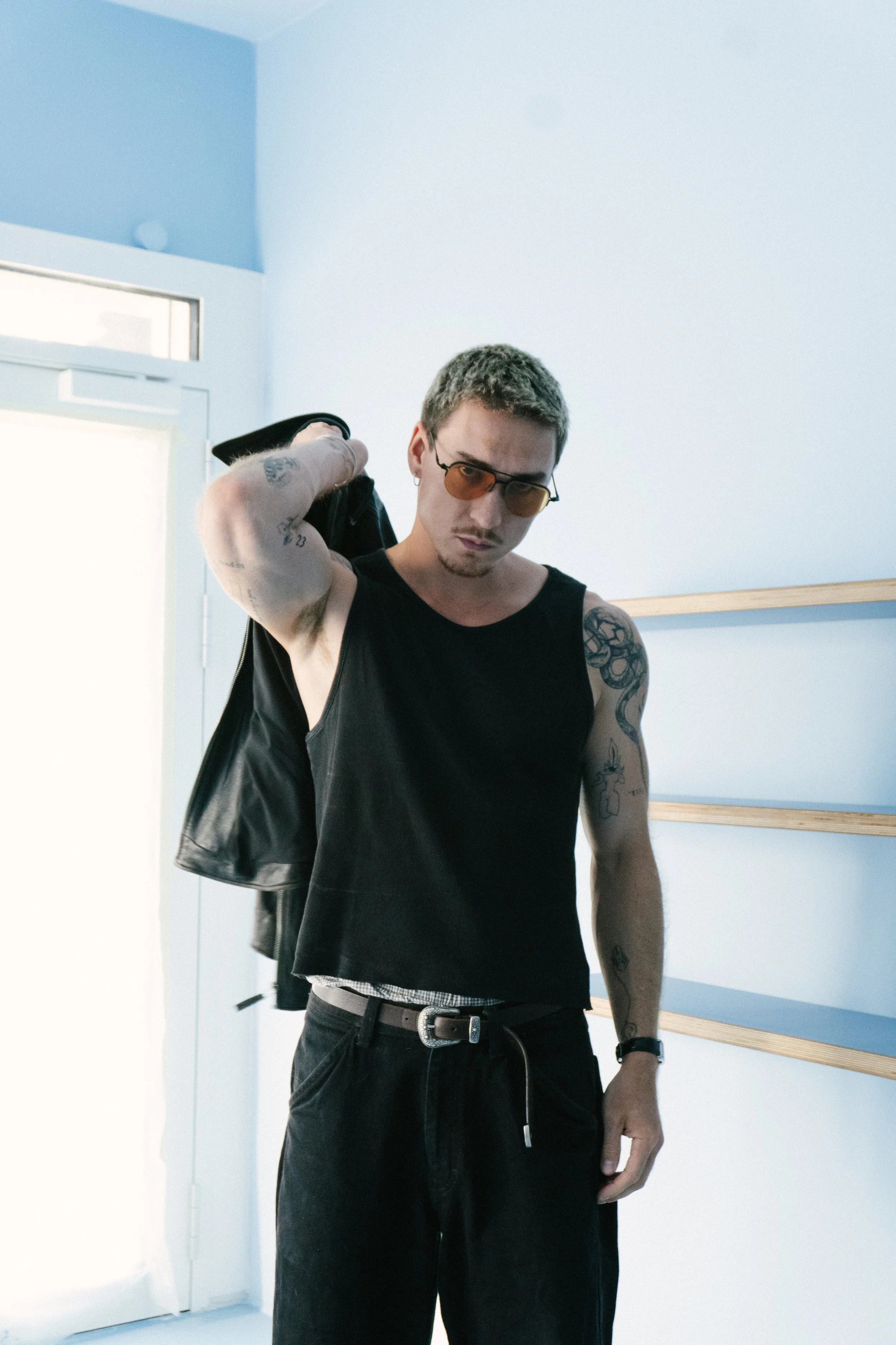 Junge Männer mit Tattoos in moderner Kleidung in einem Raum mit weißen Wänden und Holzregalen.