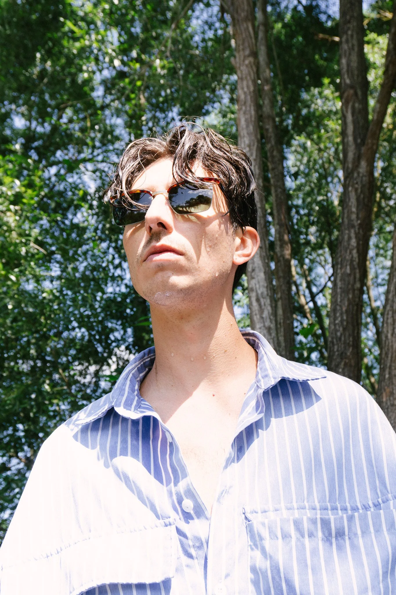 Junge Mann mit schwarzen Haaren und Sonnenbrille in einem blauen gestreiften Hemd in der Natur, hinter Bäume und Blätter