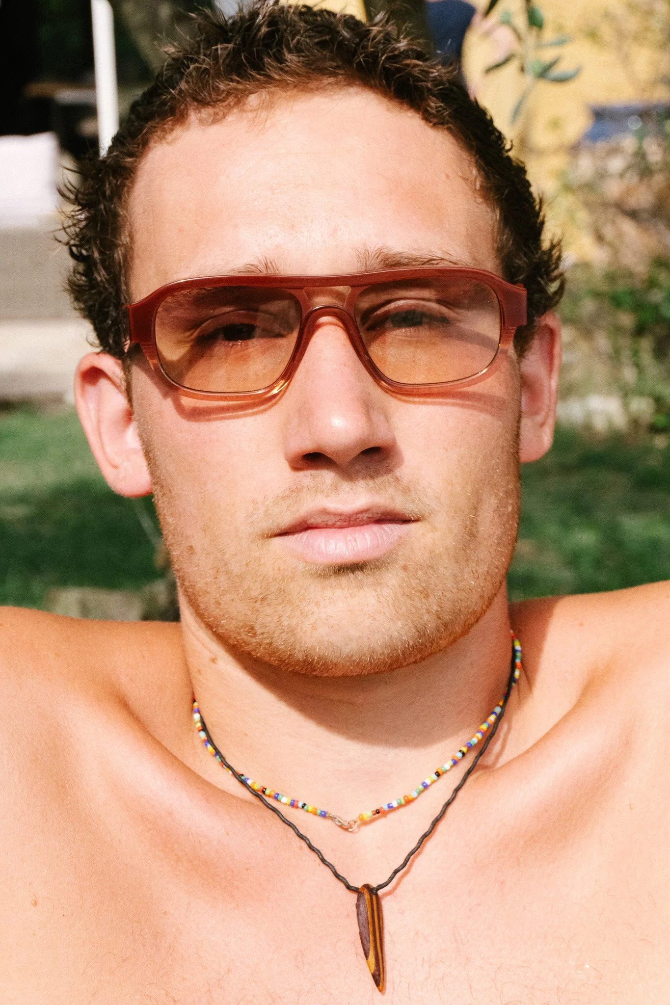 Junger Mann mit kurzen, dunklen, lockigen Haaren tragt rote Sonnenbrille und mehrere bunte Halsketten, steht im Freien bei Sonnenlicht.