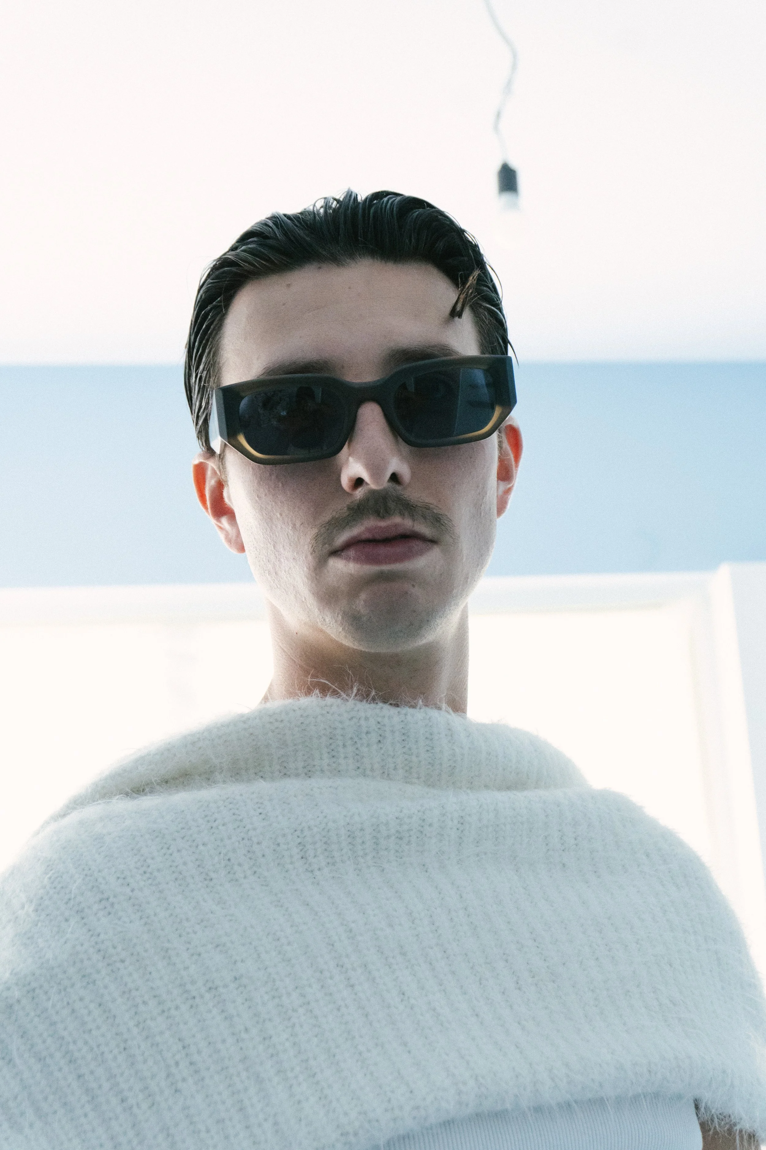 Nahaufnahme eines Mannes mit schwarzen Haaren, Sonnenbrille und weißem Pullover, im Vordergrund. Im Hintergrund blauer Himmel mit einer eingeschalteten Lampe an einer Leitung.