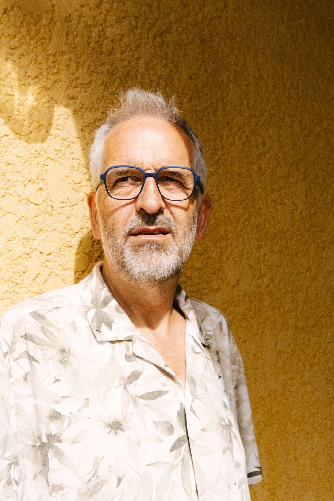 Ein älterer Mann mit grauem Haar und Bart, trägt blaue Brillen und ein sommerliches Hemd mit Blumenmuster, steht vor einer gelben Wand.