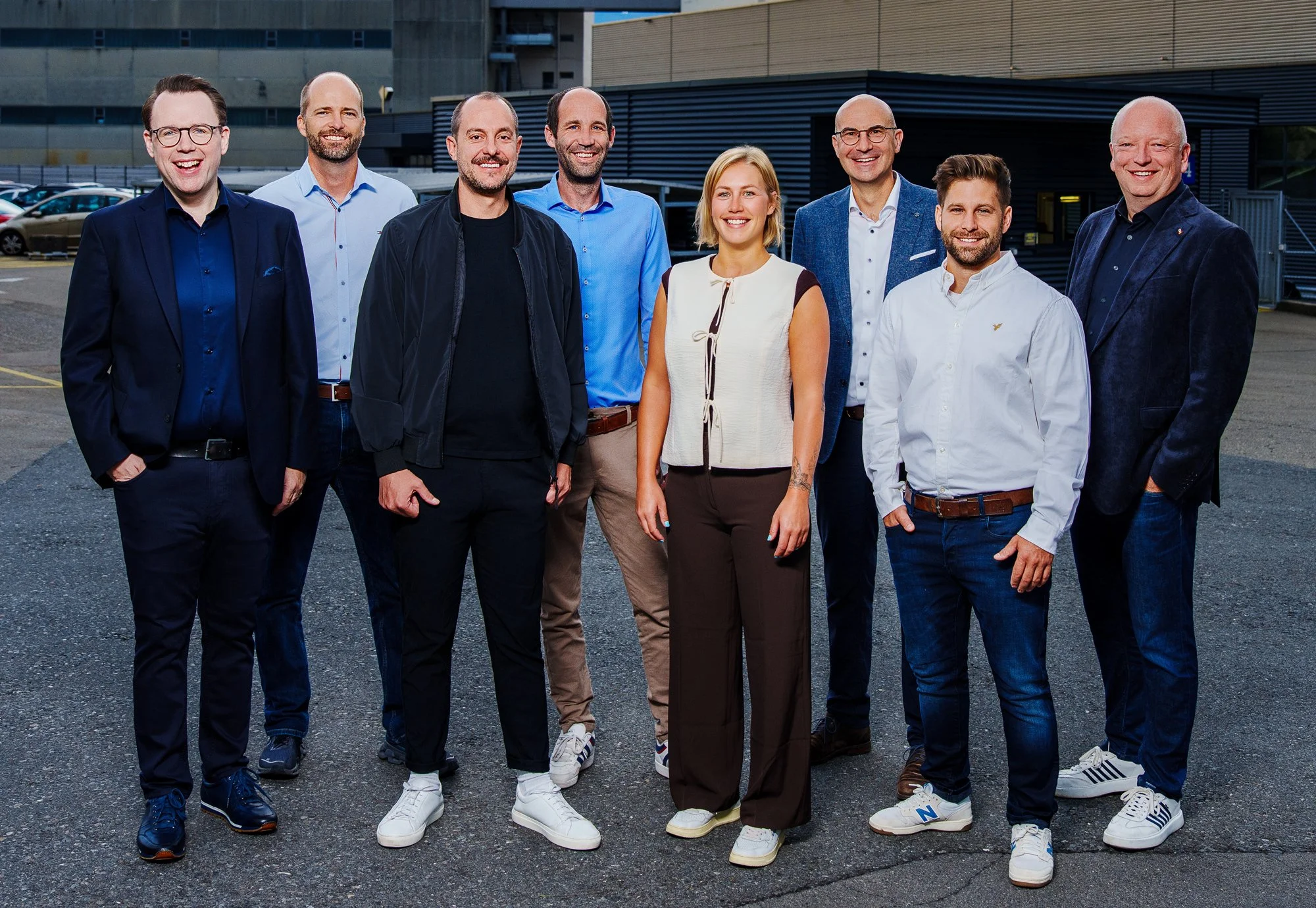 Gruppe von acht professionell gekleideten Menschen, die draußen vor einem modernen Gebäude stehen und lächeln