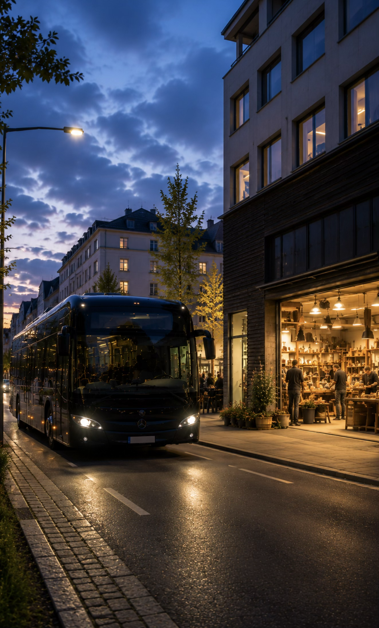Abendkulisse in einer Stadt mit einem beleuchteten Bus, einem offen stehenden Laden mit warmem Licht und Menschen, die darin einkaufen oder sich unterhalten, vor einer modernen Gebäudefront bei bewölktem Himmel.