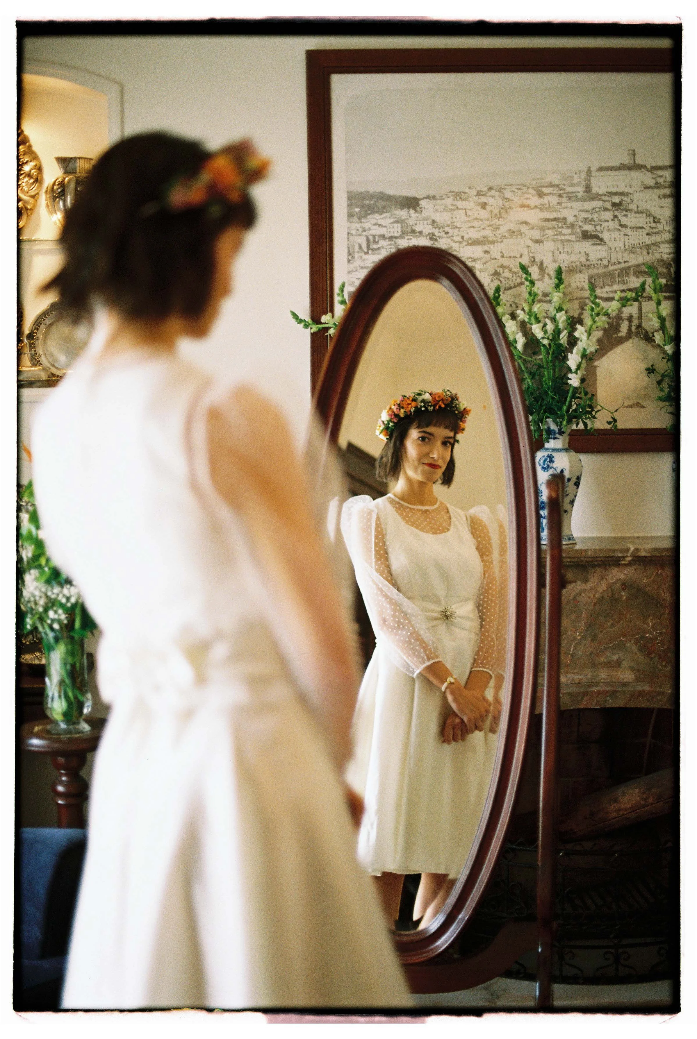 Bride looking at her reflection in an oval mirror, wearing a flower crown in a room decorated with plants and a city painting on the wall, photographed at a wedding in Portugal.