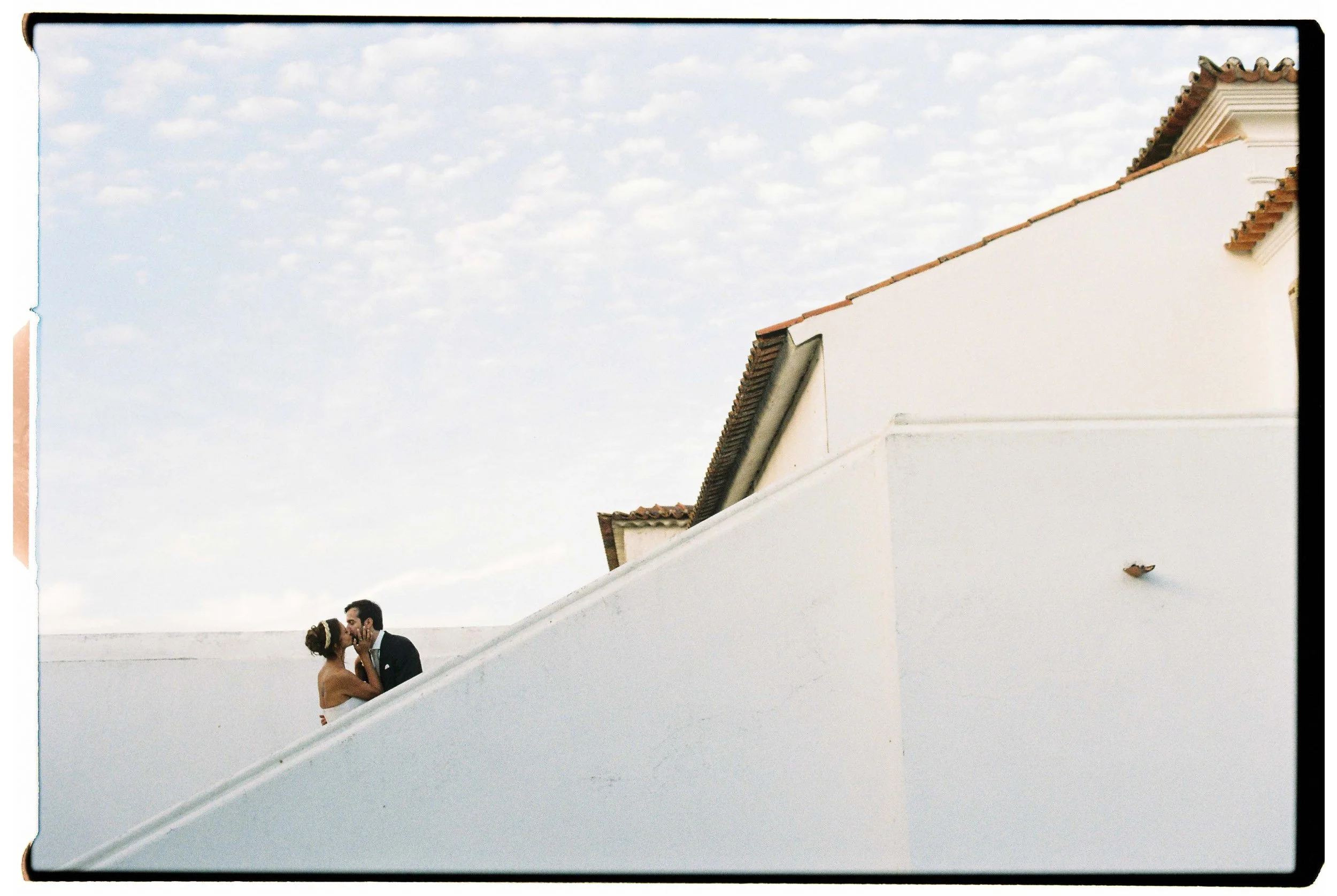 Wedding couple standing at the top of a white staircase, with a red-tiled roof and soft white clouds in the sky behind them, photographed at a wedding in Portugal.