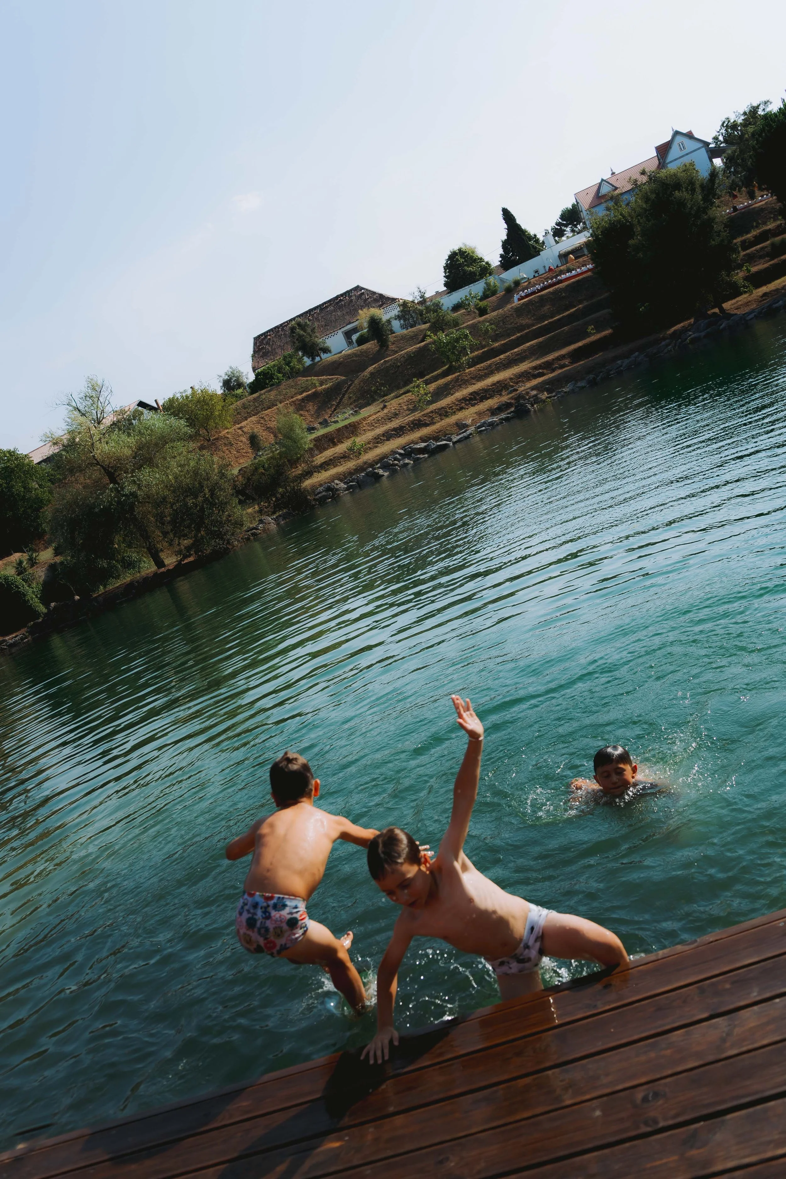 Children playing in the water with adults, surrounded by trees and houses in the background, photographed at a wedding in Portugal.
