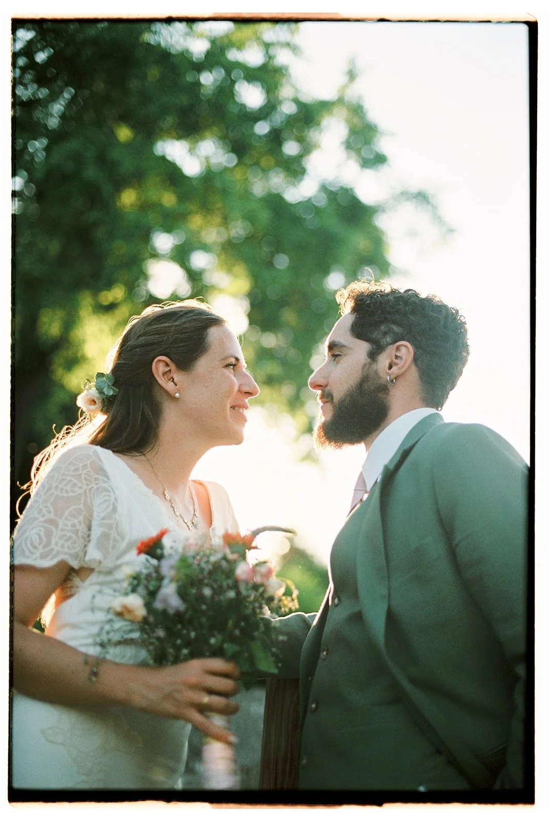 Bride and groom smiling at each other outdoors, surrounded by greenery and natural evening light, holding a bouquet of flowers, photographed at a wedding in France.