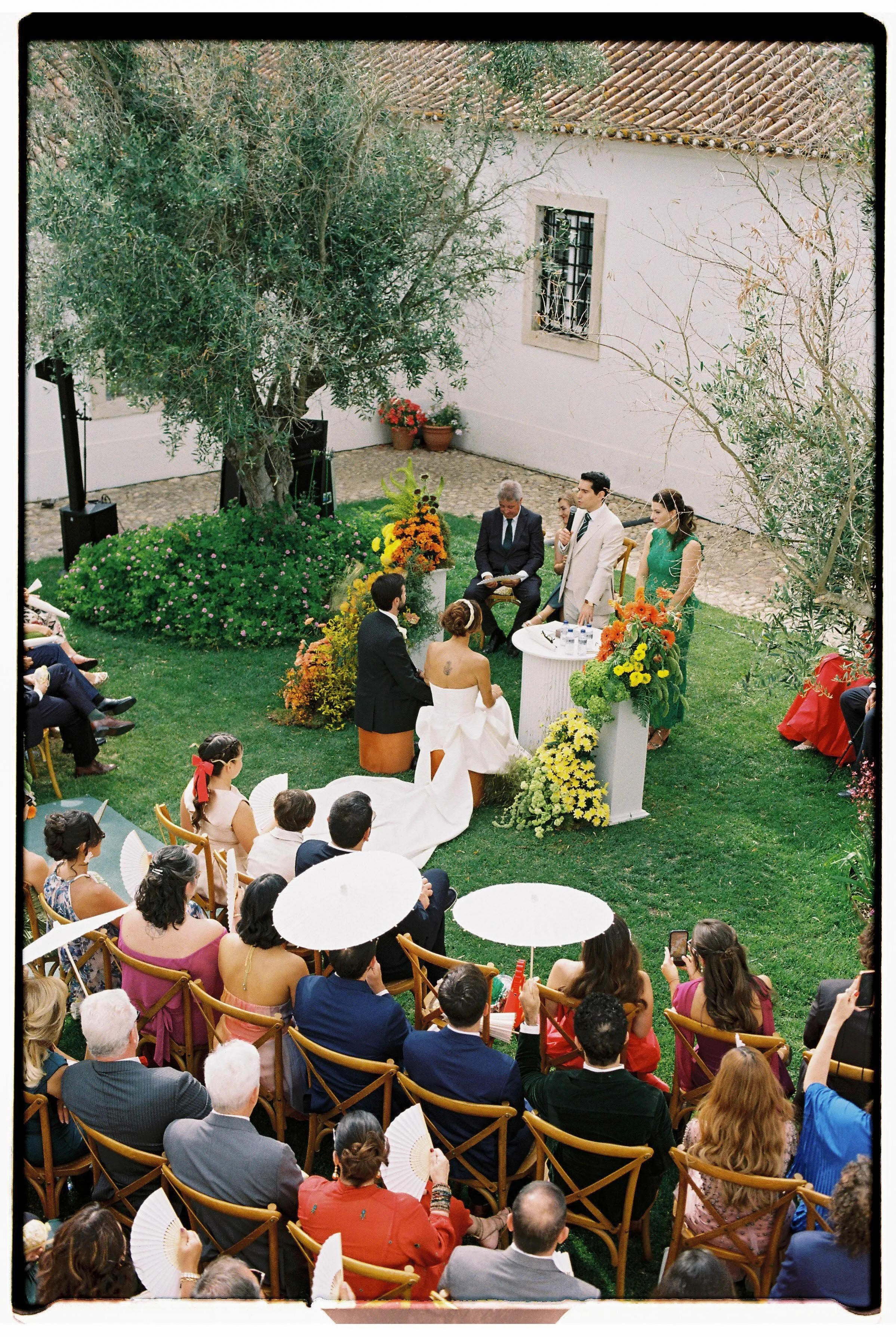 Outdoor wedding ceremony with guests seated on wooden chairs, surrounded by trees and greenery, with the couple seen from behind at the altar, photographed at a wedding in Portugal.