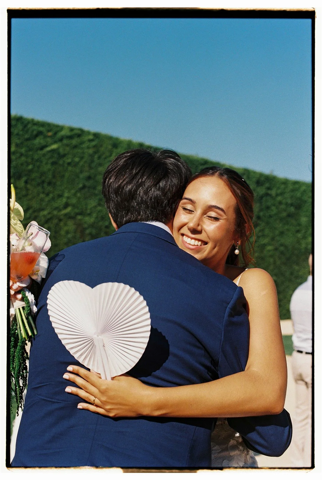 Couple embracing as the bride smiles and holds a paper heart, surrounded by greenery under a blue sky, photographed at a wedding in Portugal.