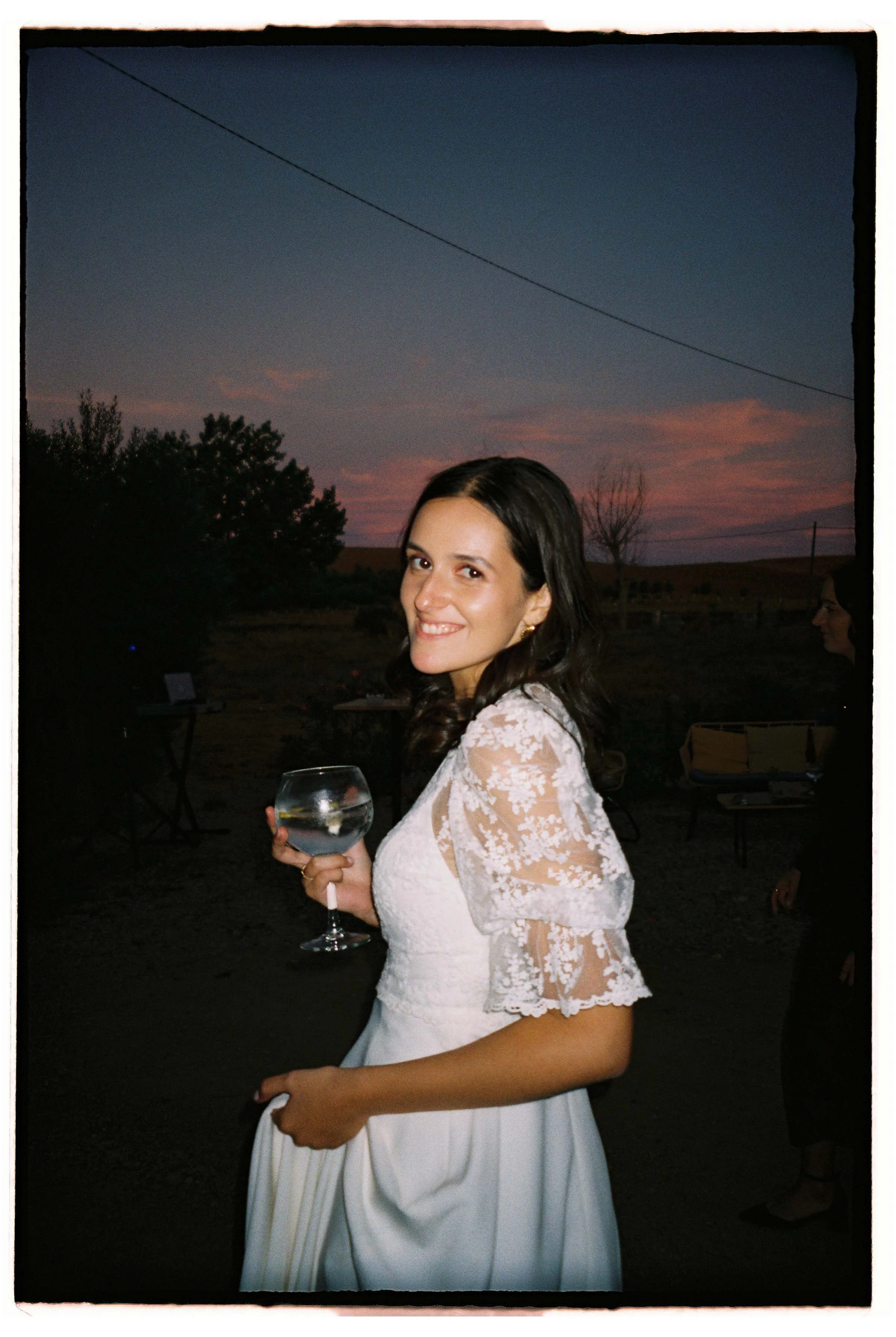 Bride smiling in her wedding dress, holding a glass of wine outdoors at sunset, with a colourful sky in the background, photographed at a wedding in Alentejo, Portugal.