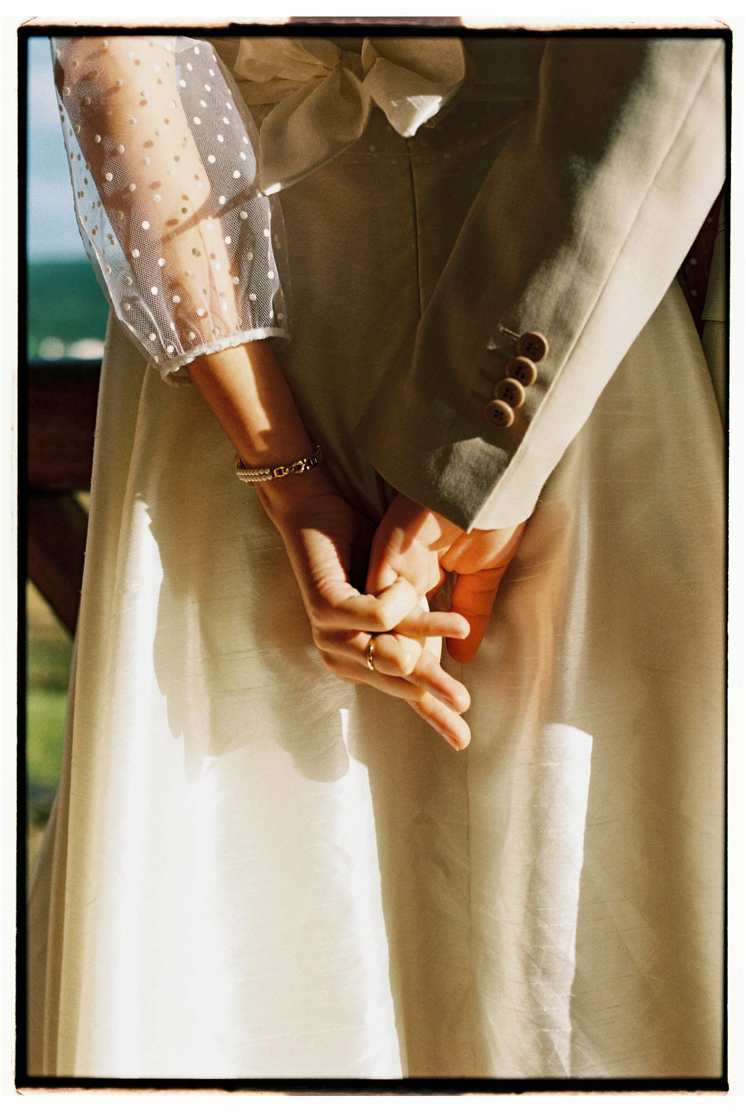 Intertwined hands in formal attire, with a beige blazer and sleeve buttons, against a green field outdoors, photographed at a wedding in Portugal.